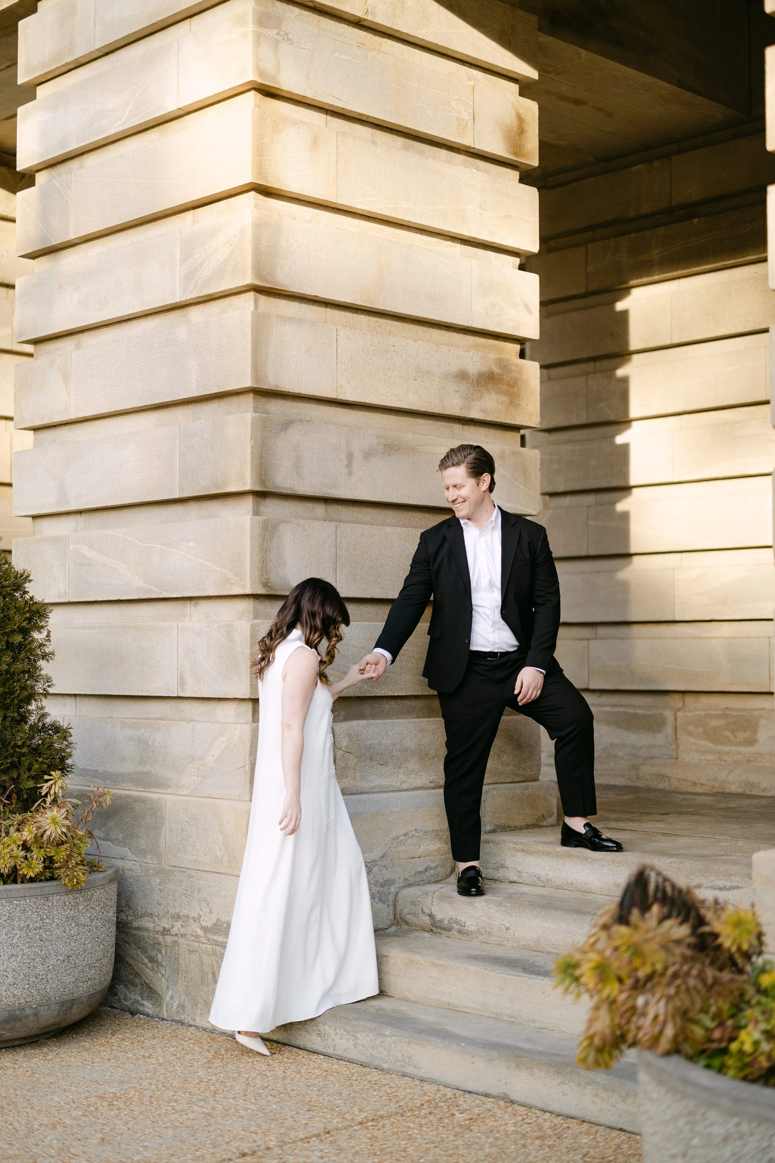 A man in a black suit helping a woman in a white dress down some stone stairs outside a large stone building.