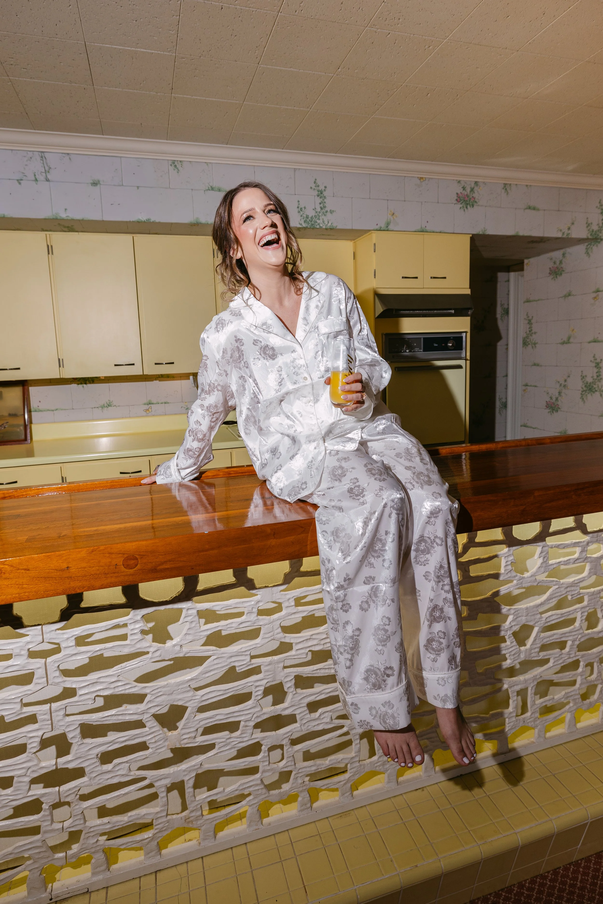 A woman in white floral pajamas sitting on a wooden kitchen countertop, holding a glass of orange juice, smiling and laughing.
