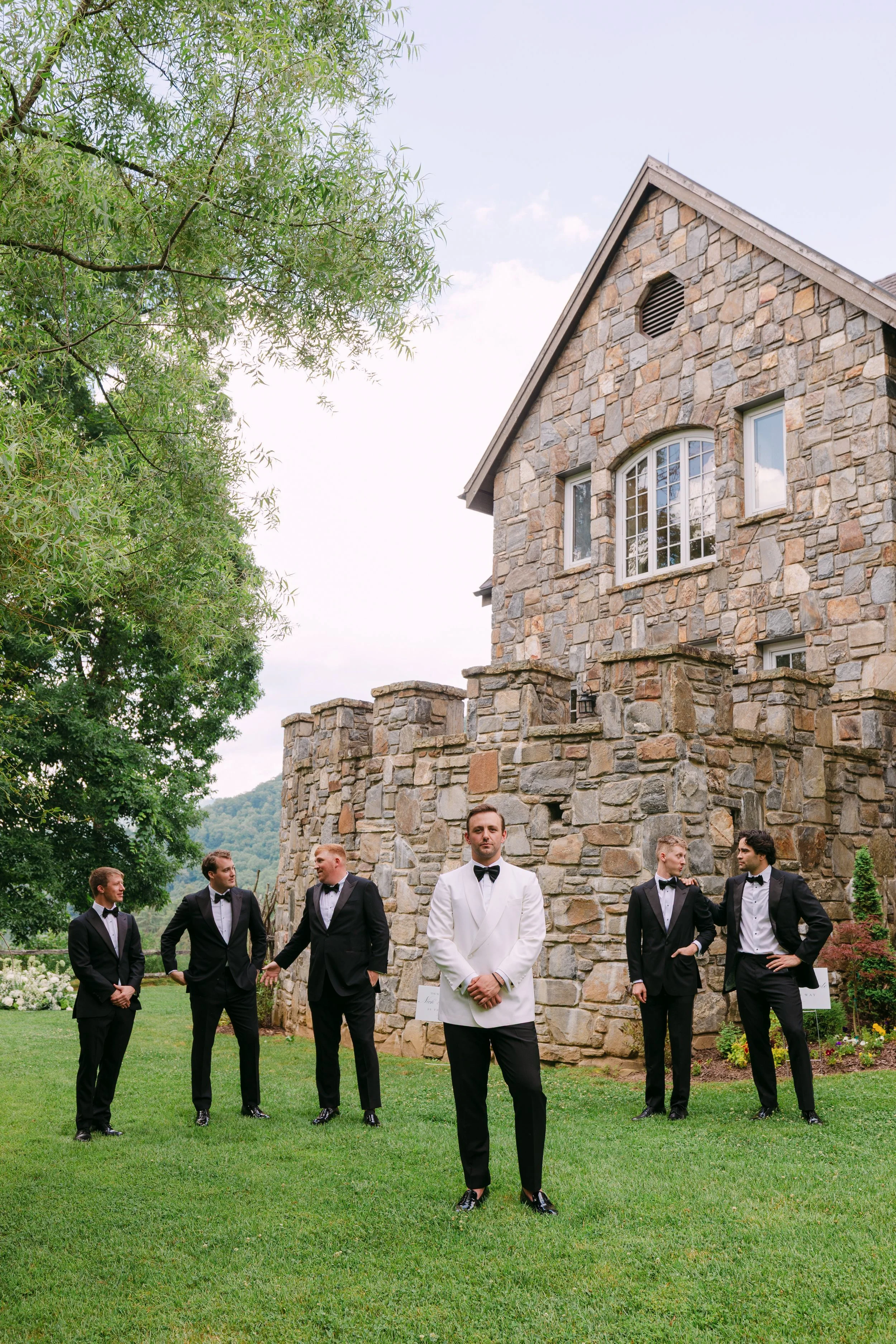 Six men in formal tuxedos standing on lawn in front of stone mansion, one in white tuxedo jacket in center, others in black tuxedos with bow ties, with green trees and cloudy sky in background.