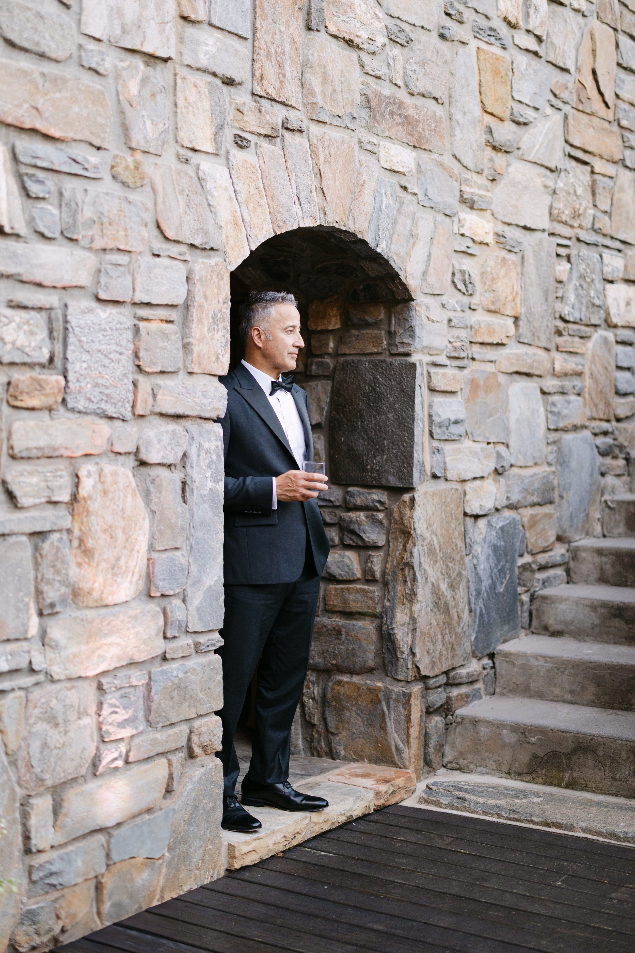 A man in a tuxedo holding a glass of drink, standing on a small stone platform by a stone wall and staircase.