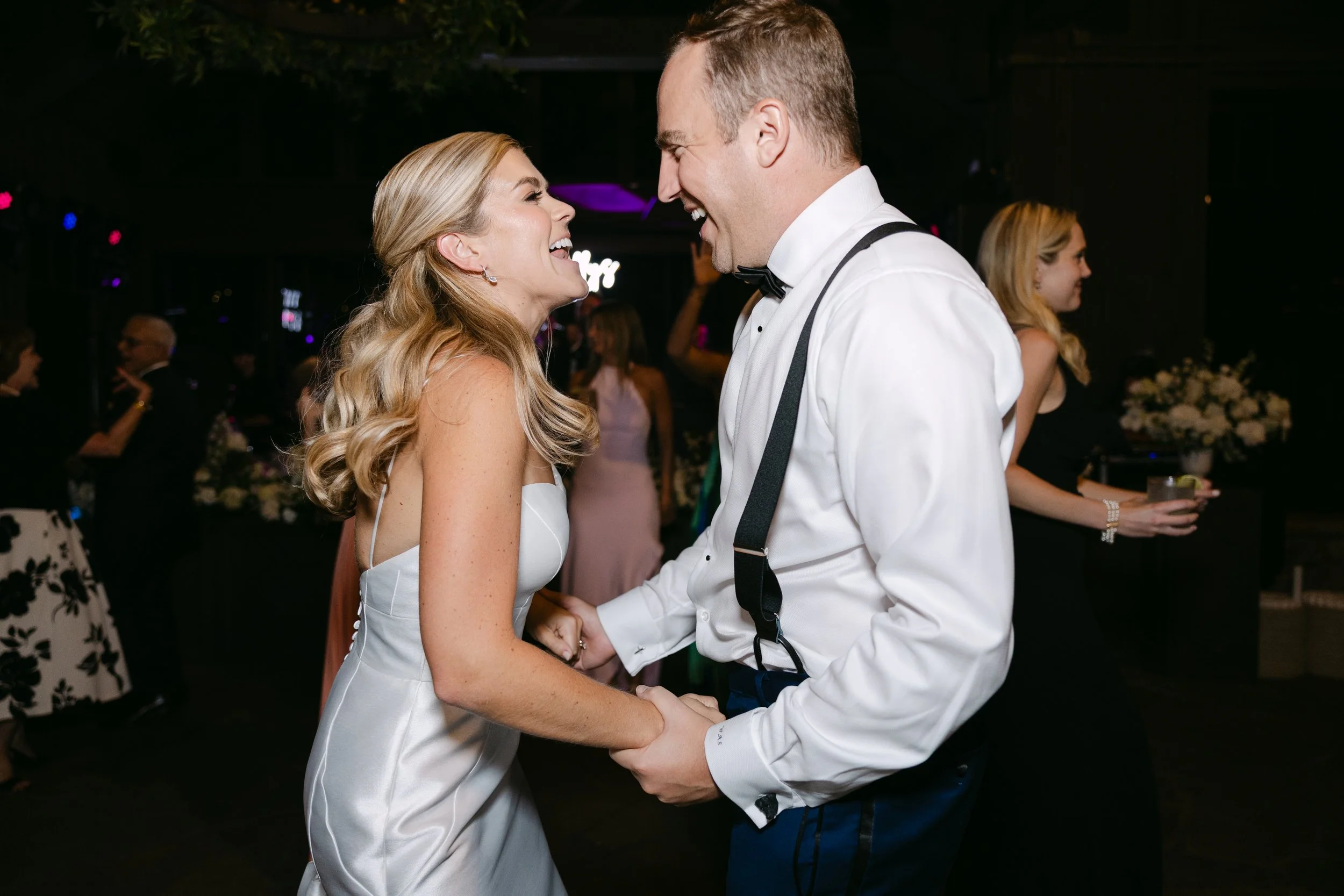 A bride and groom are smiling and holding hands while dancing at their wedding reception. The bride has long blonde hair and is wearing a white wedding dress, and the groom is wearing a white shirt with suspenders and a black bow tie. Other guests ar