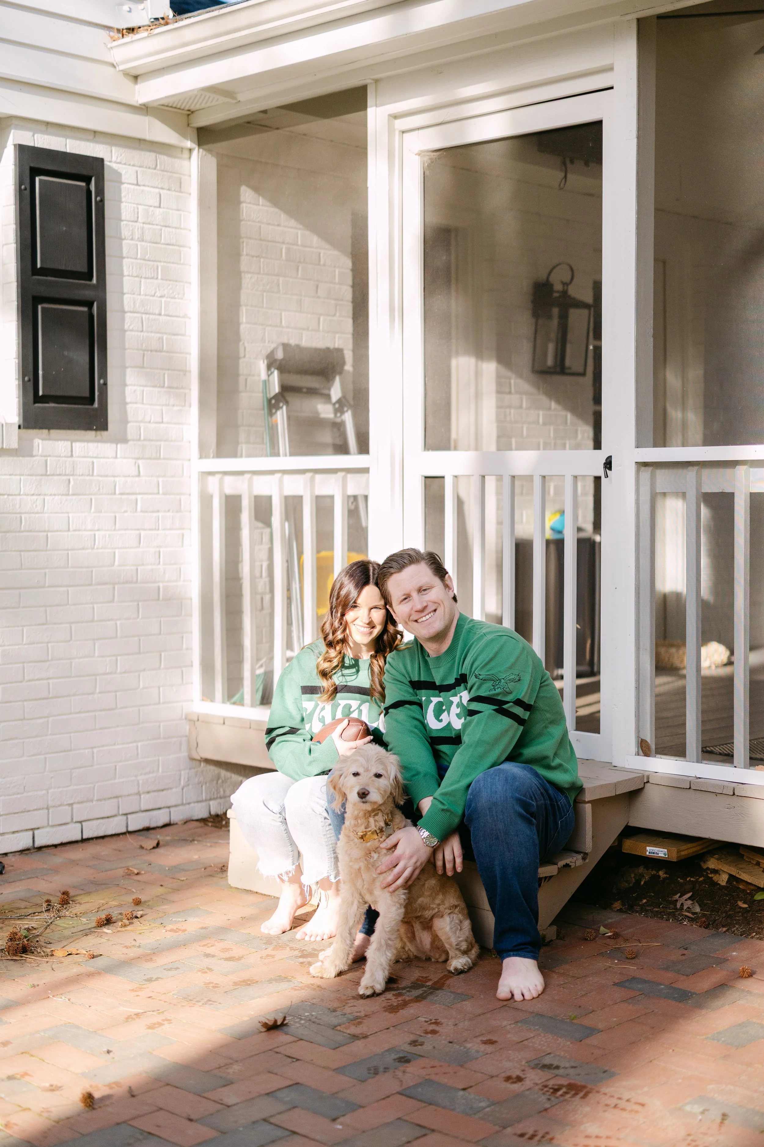 A smiling couple sitting outside on brick steps with their light-colored puppy, in front of a porch with white railing and brick wall.