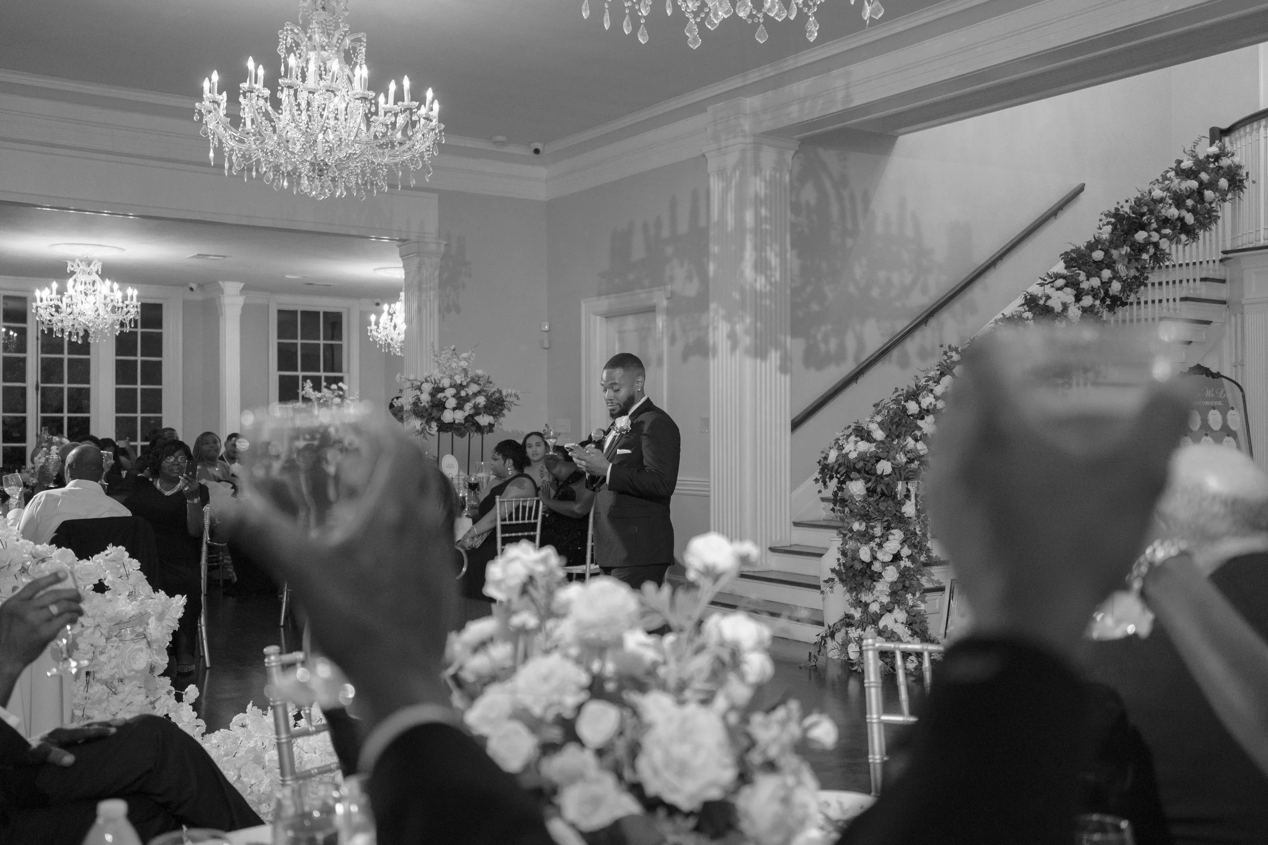 A formal event with guests seated at decorated tables, chandeliers hanging from the ceiling, and a man in a suit standing near a floral arch.