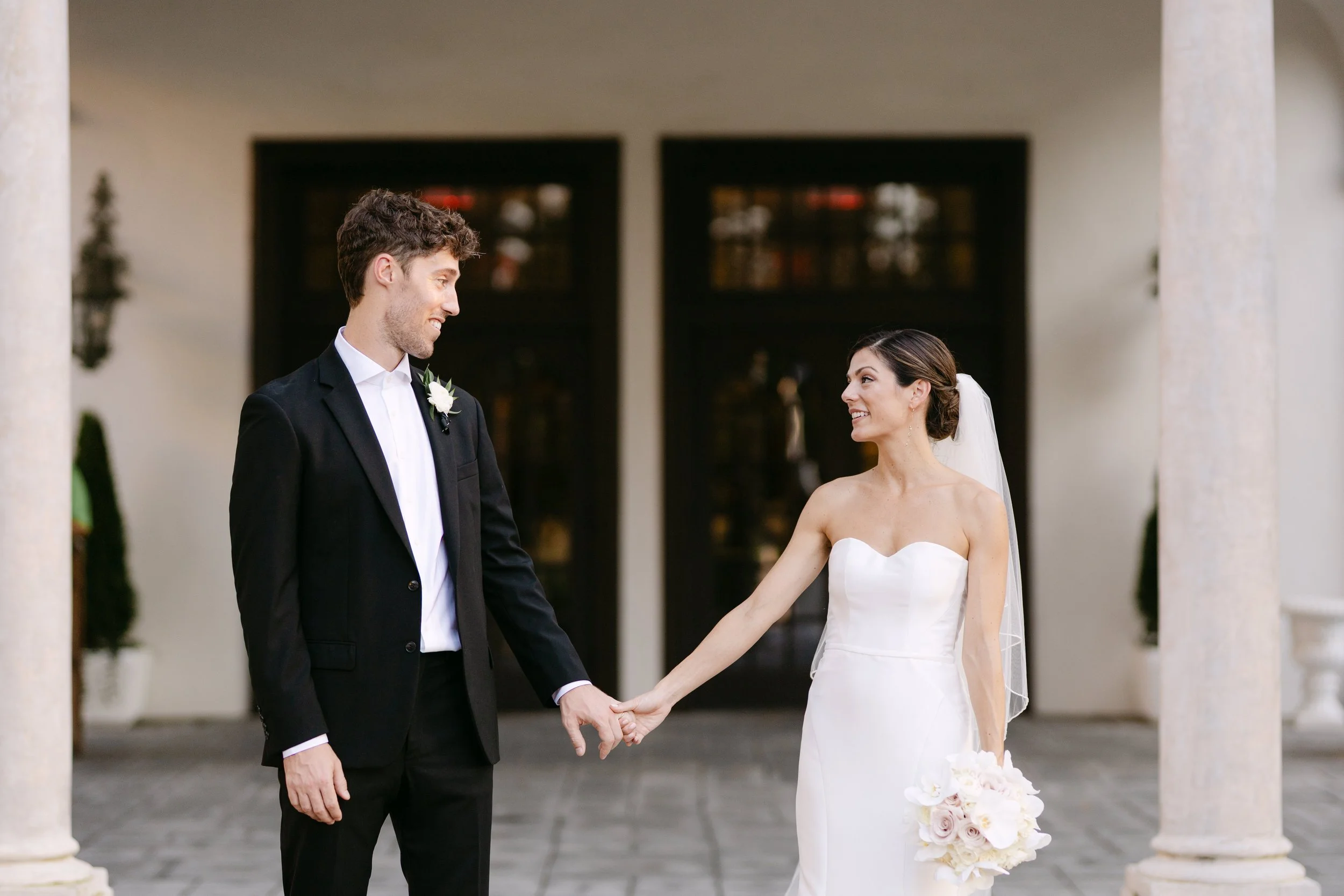 A bride and groom holding hands outside a building with columns, smiling at each other. The bride is in a strapless white wedding dress and veil, holding a bouquet of pale pink and white flowers. The groom is in a black suit with a white shirt and bo