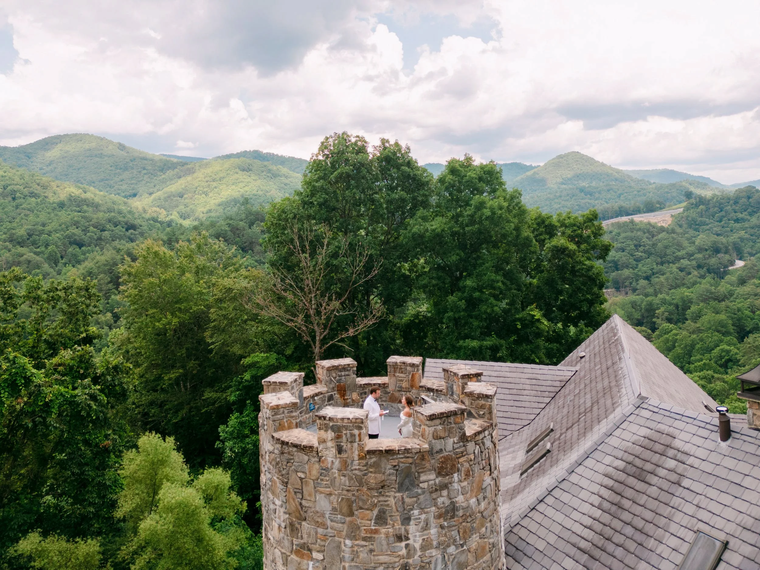 A stone tower with a wedding ceremony occurring on top, surrounded by green trees and mountain hills in the background.