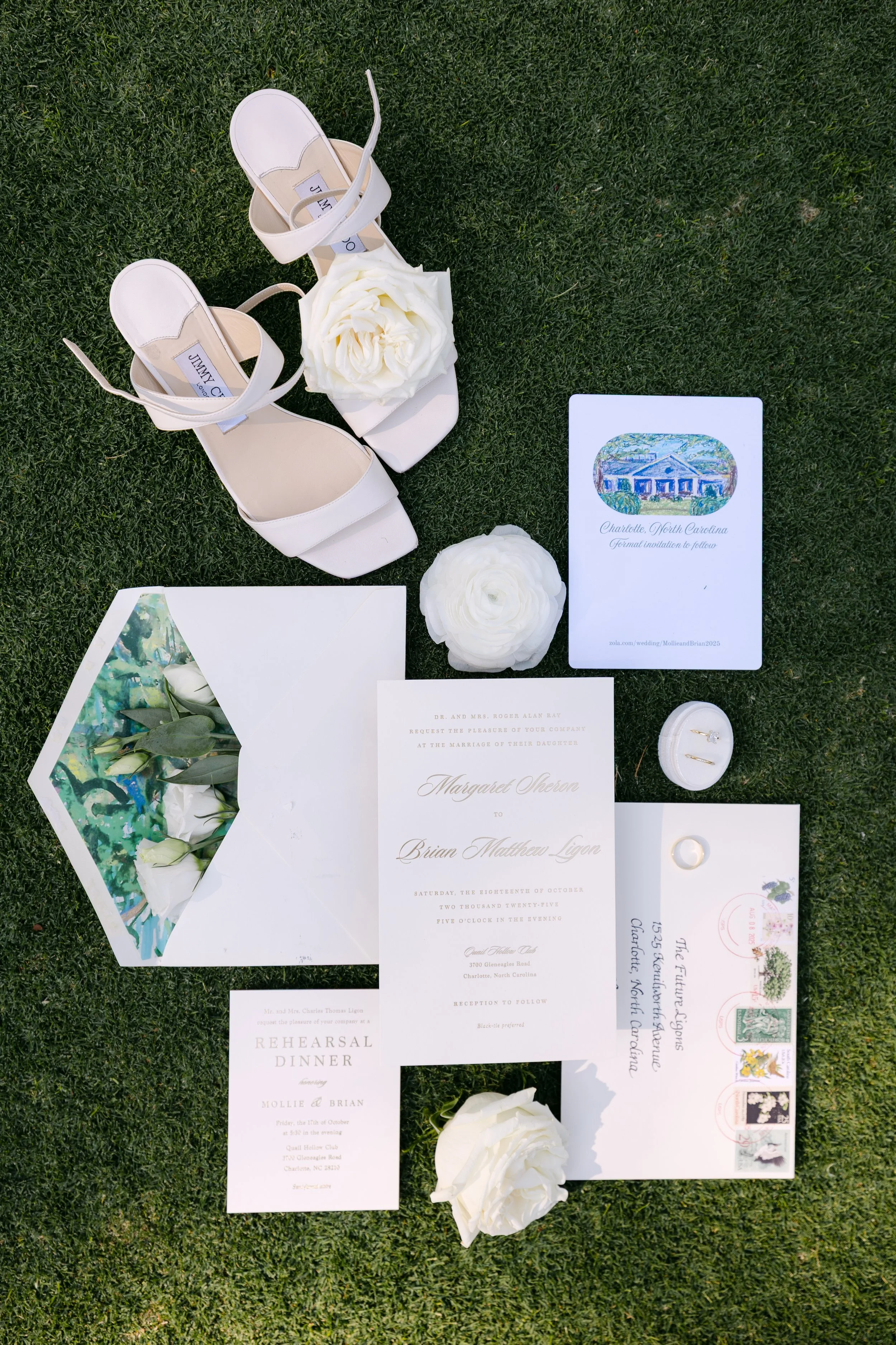 White high-heeled sandals with flower embellishments and a collection of wedding invitations, envelopes, flowers, and rings arranged on grass.