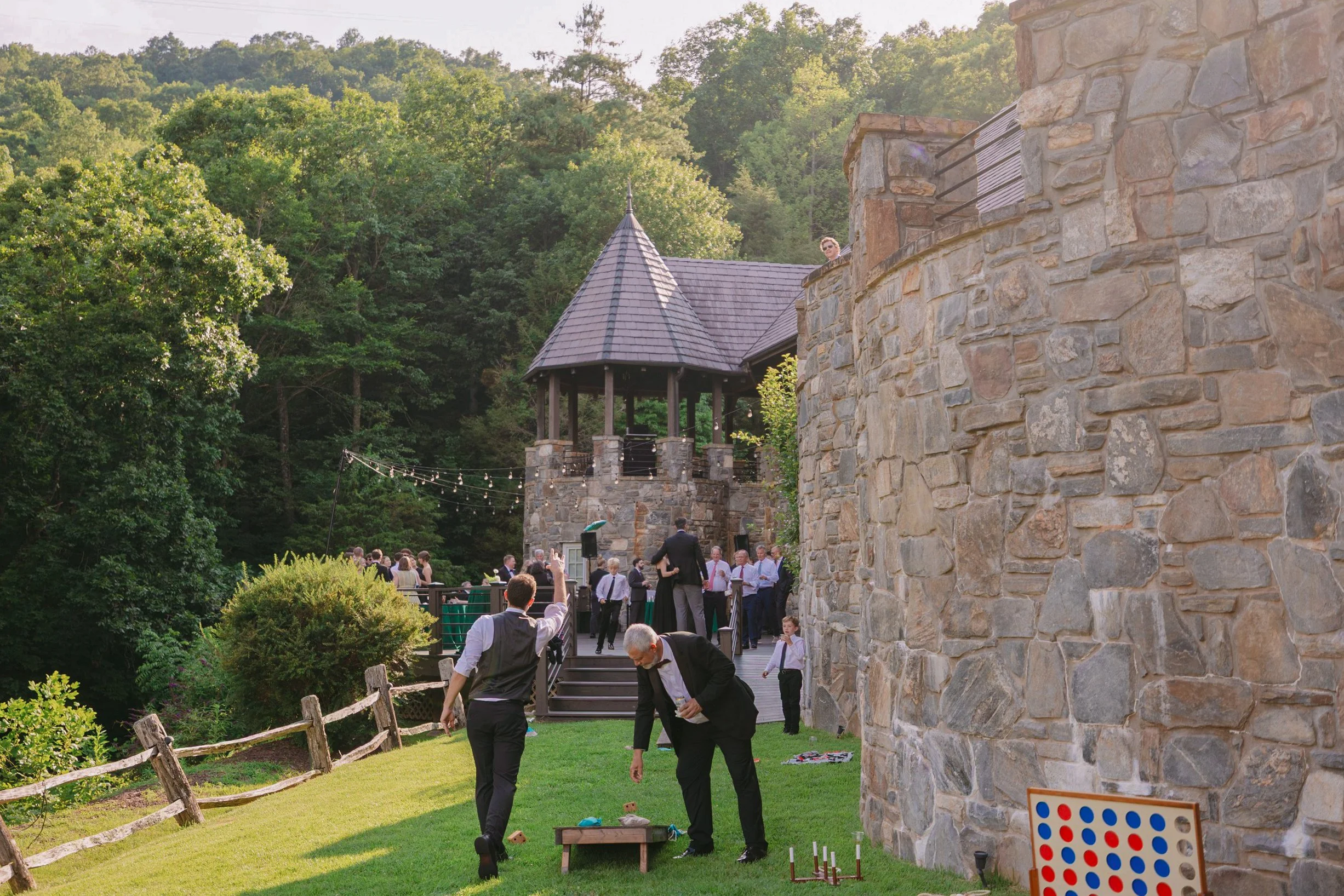 People gathered outdoors on a wooden deck, socializing and enjoying the event, with some standing near a stone tower and others across the grassy area playing games like giant Jenga and Connect Four, surrounded by lush green trees.