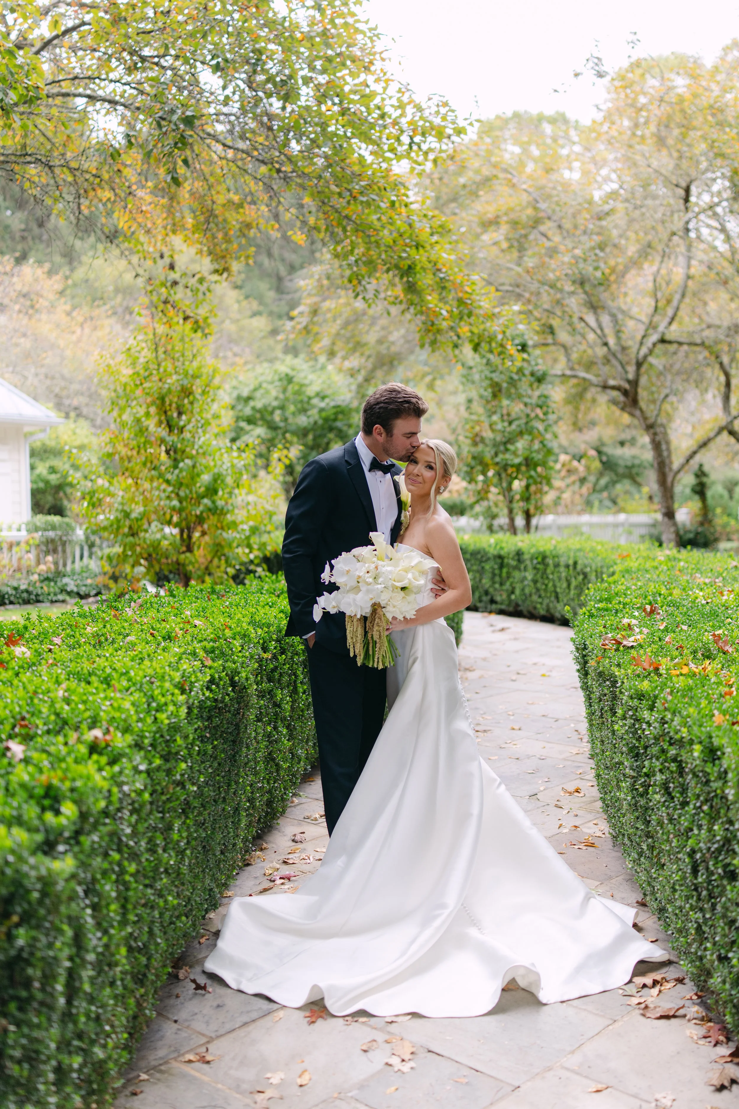 Bride and groom embracing on a garden path, with the bride holding a white floral bouquet.