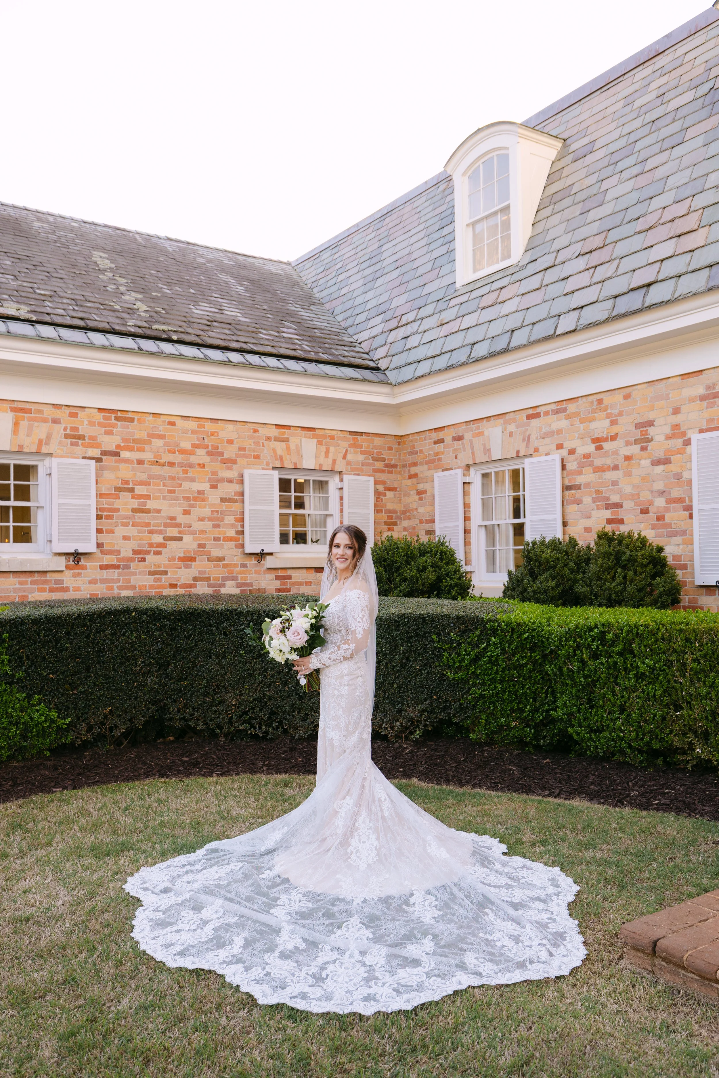 A bride in a lace wedding dress holding a bouquet of flowers, standing on a lawn in front of a brick house with white window shutters and green bushes.