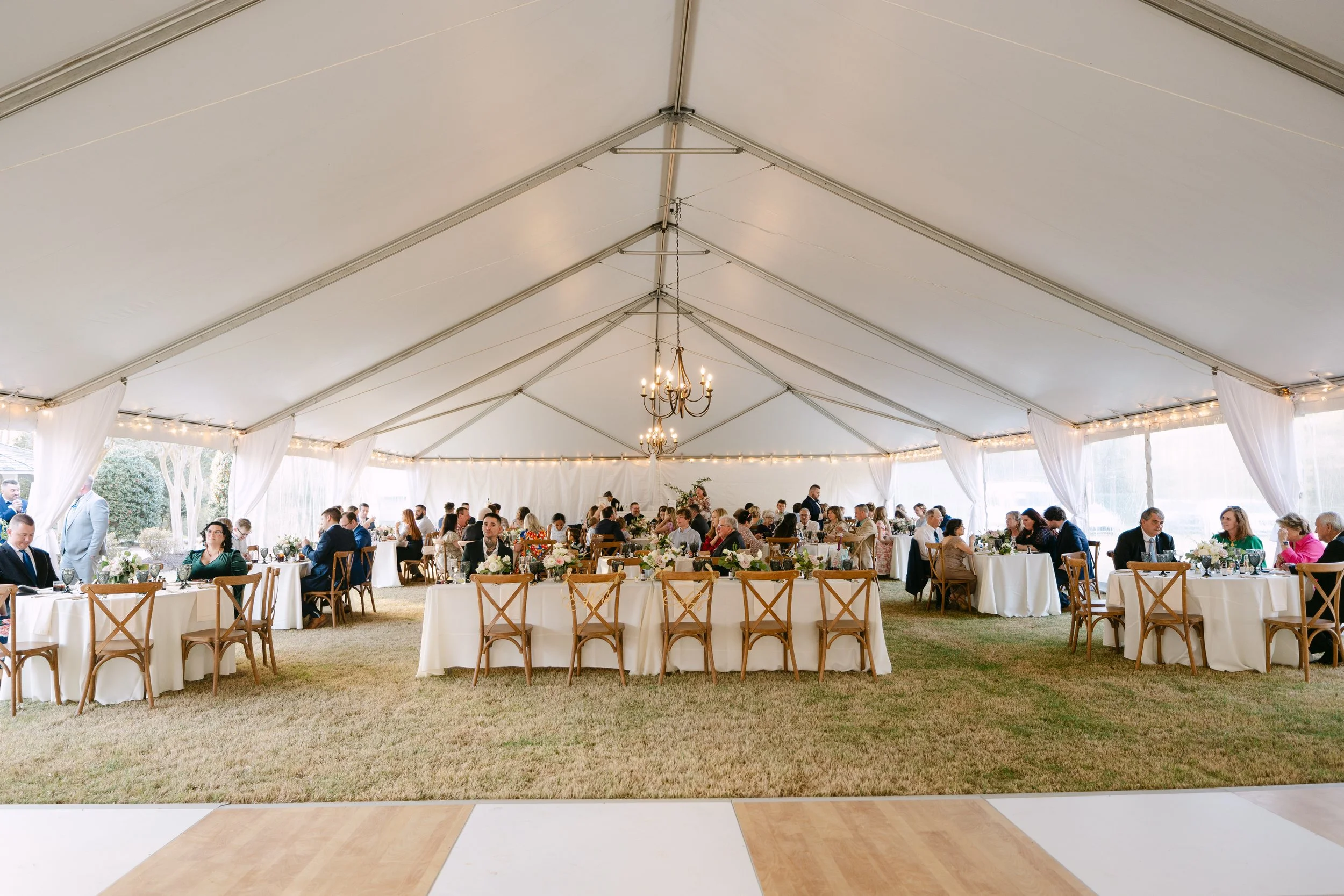 A large wedding reception tent with round tables decorated with white tablecloths and floral centerpieces. Guests are seated and dining, and the tent is lit with chandeliers and string lights, with an open view of the outdoors.