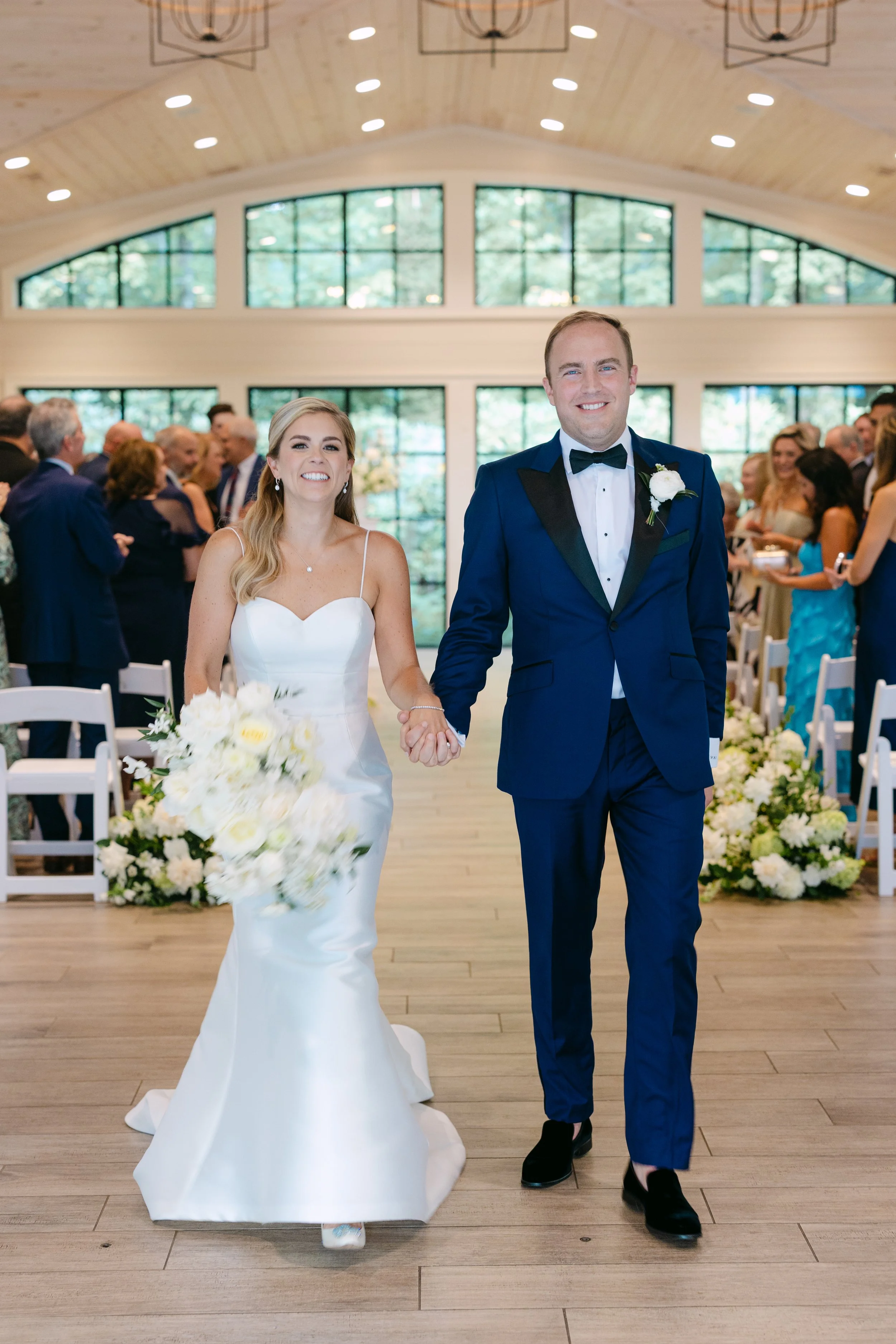A bride and groom walking down the aisle during their wedding ceremony, holding hands and smiling, with family and friends seated and standing on either side. The bride is wearing a white wedding gown and holding a large bouquet of white flowers, whi