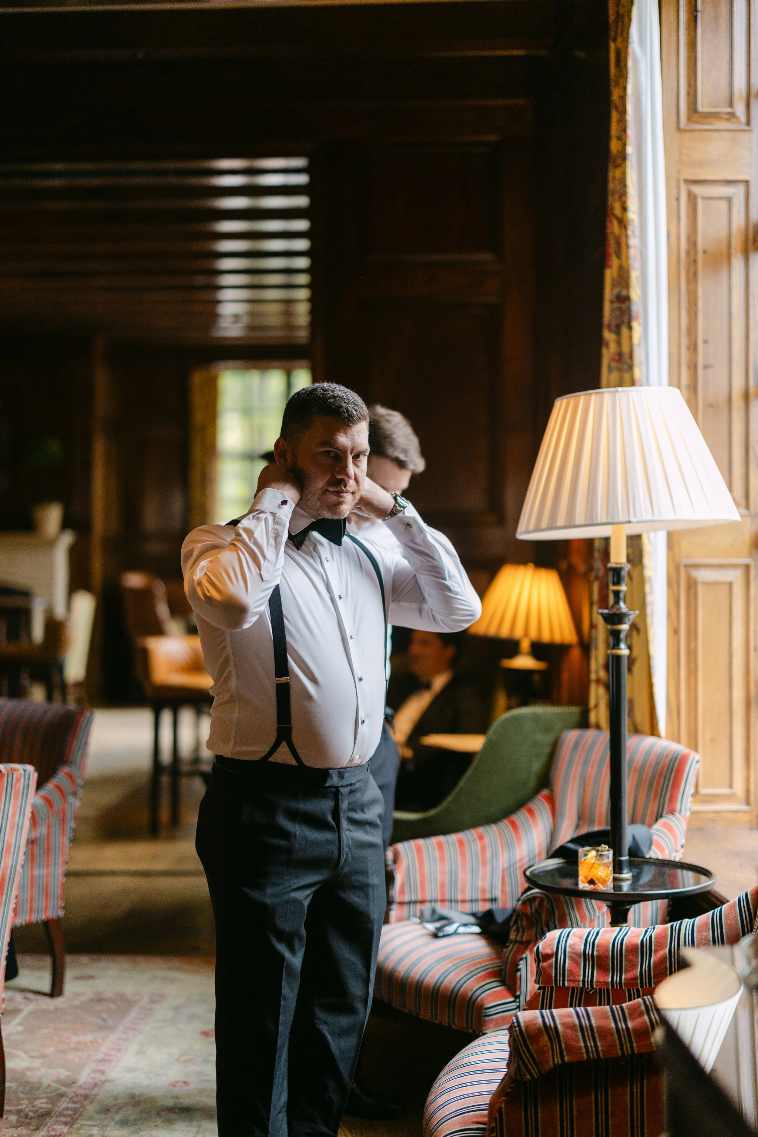 A man adjusting his suspenders in a warmly lit, elegant room with wood-paneled walls, striped upholstered chairs, and table lamps. In the background, a woman is sitting at a table.