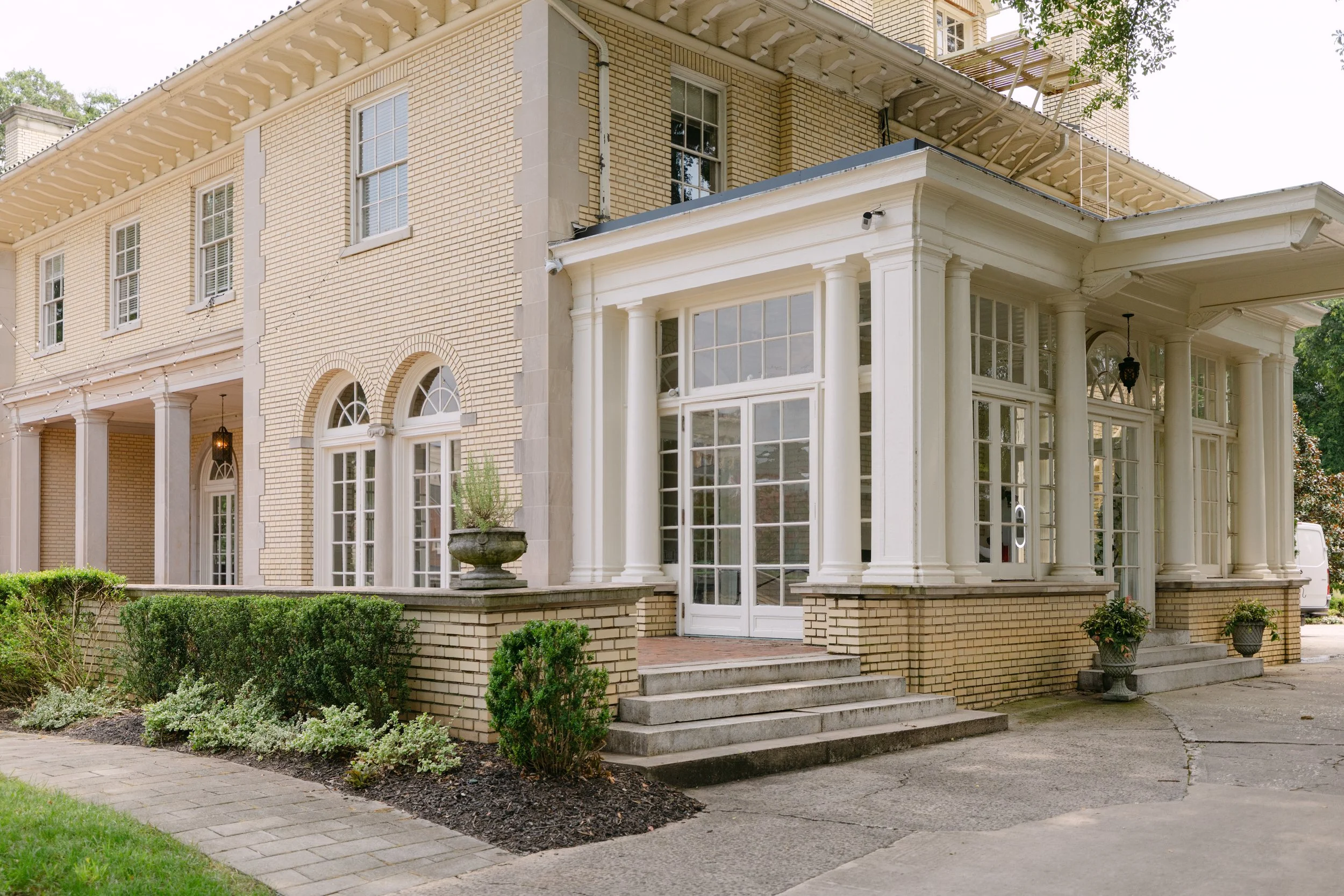 The image shows the front exterior of a large, traditional house with beige brick walls, white columns, and multiple windows. There are steps leading up to the entrance, greenery, and potted plants in front of the house.