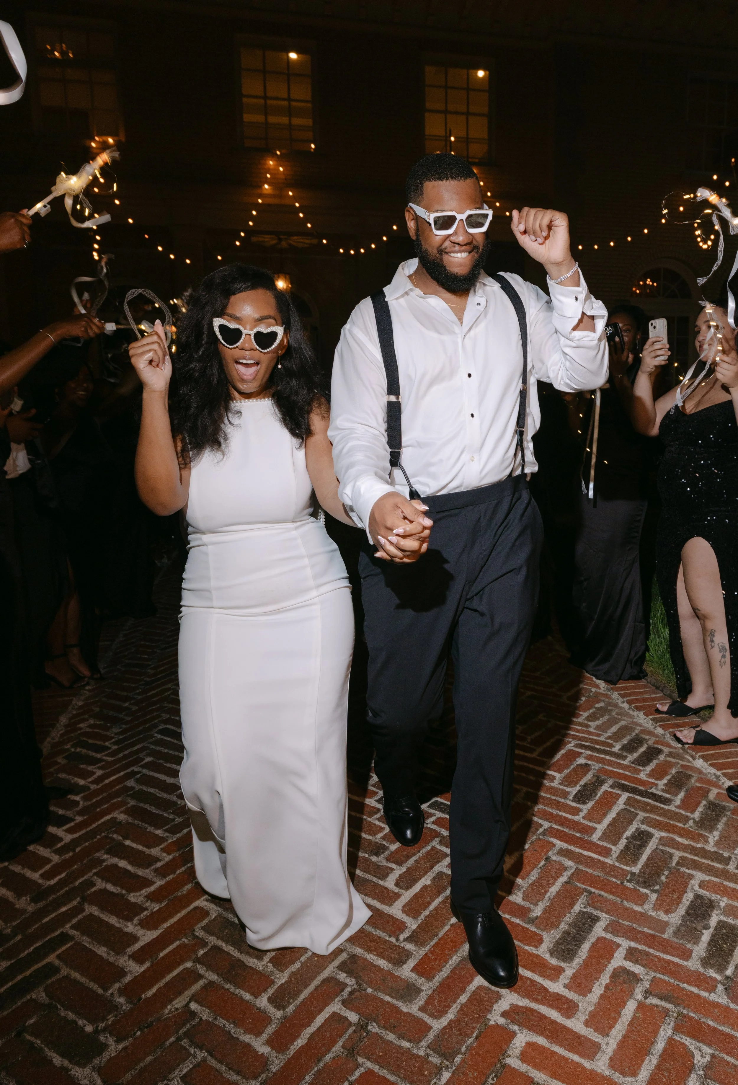 Group of people celebrating at night, holding sparklers, with fairy lights in the background. The couple in the foreground is dancing and wearing stylish glasses, dressed in formal attire.