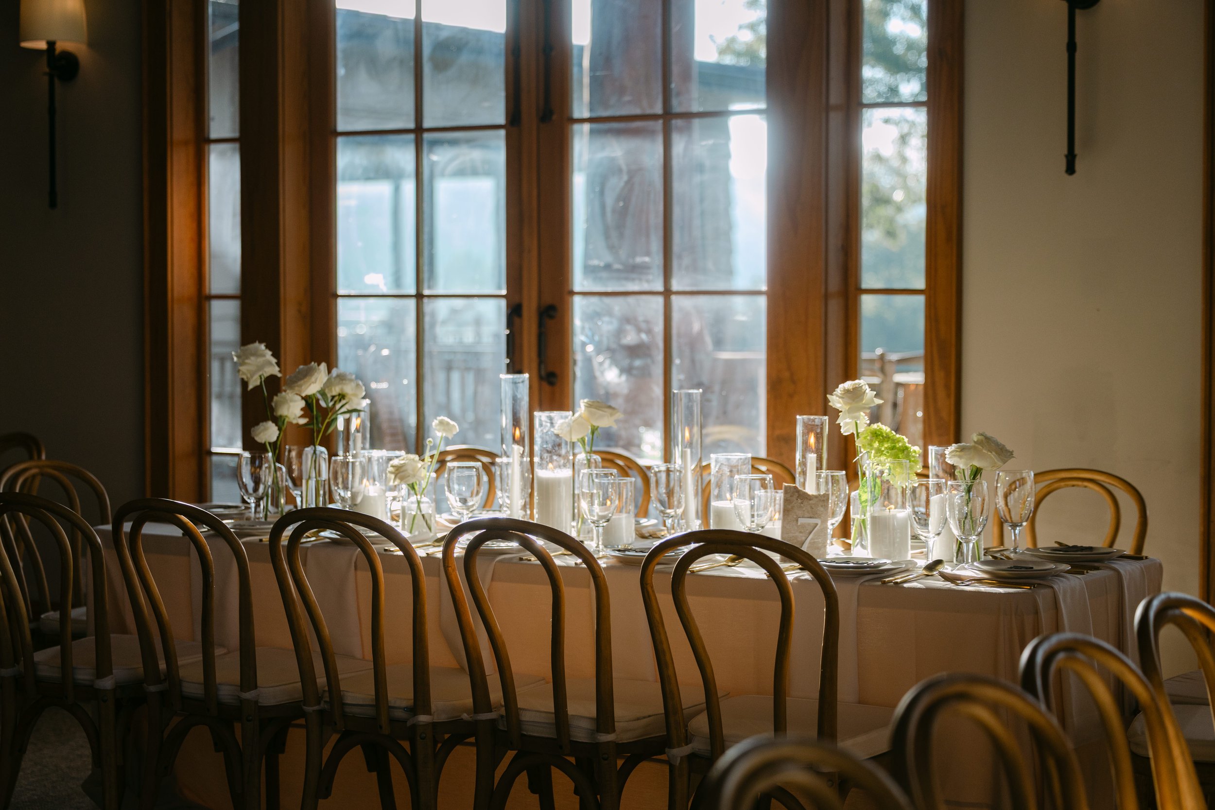 Elegant dining table set with white flowers, candles, and glassware in front of large wooden windows with sunlight streaming in.