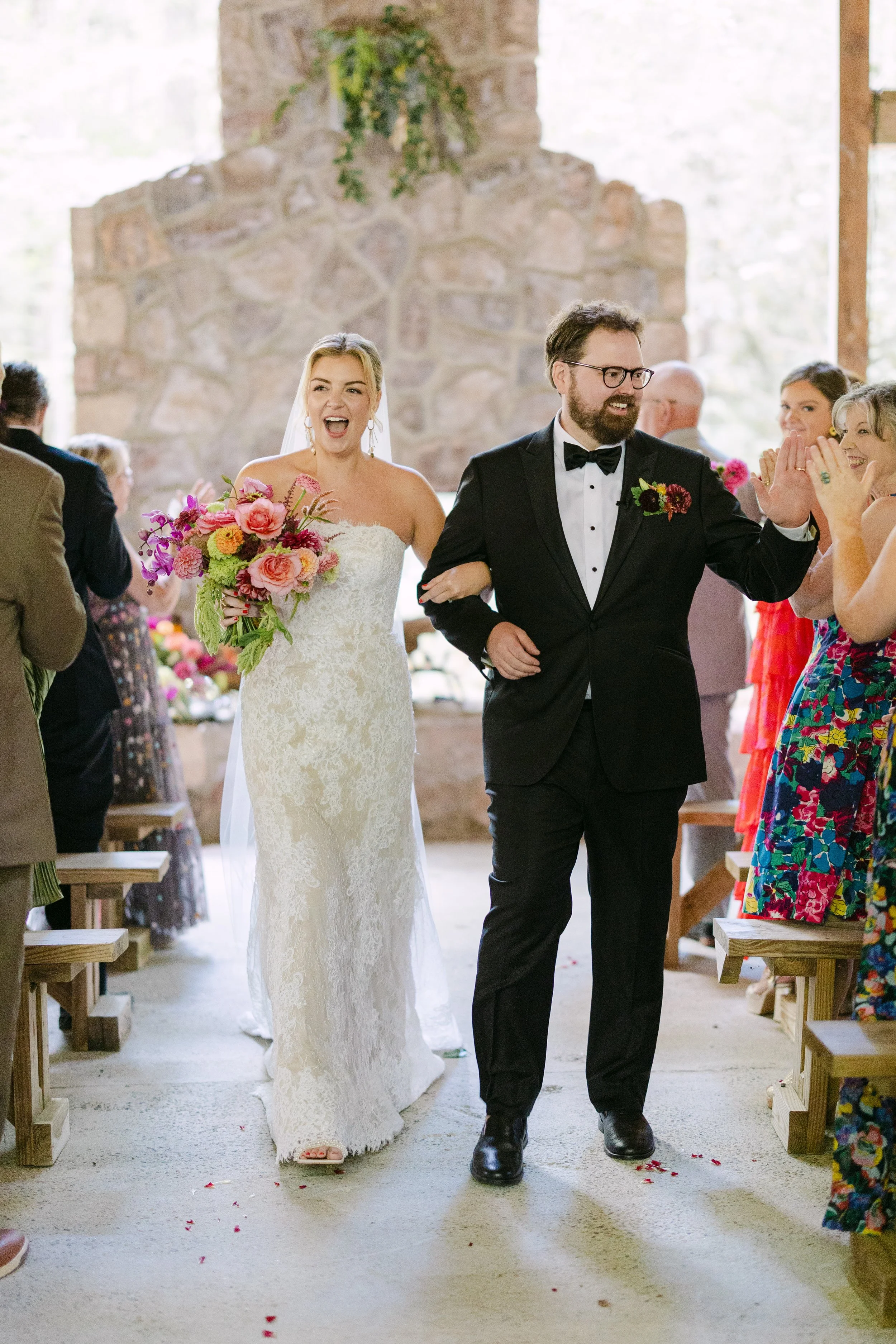 A bride and groom walking down the aisle, smiling, wedding guests clapping and celebrating inside a rustic venue with stone walls and large windows.