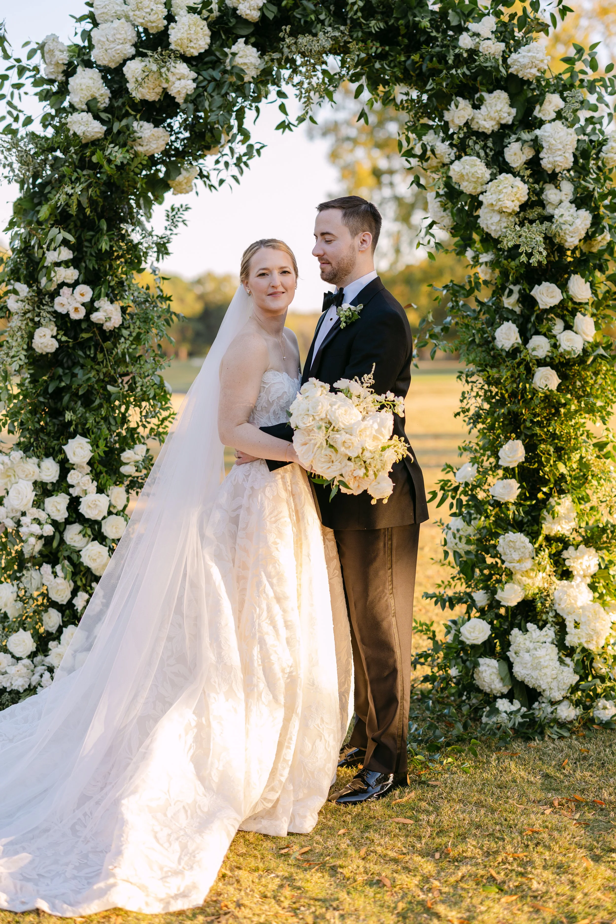 A bride and groom standing under a floral arch during their wedding, outdoors in a park, with sunlight and trees in the background. The bride wears a white wedding gown and veil, holding a bouquet, and the groom wears a black tuxedo.