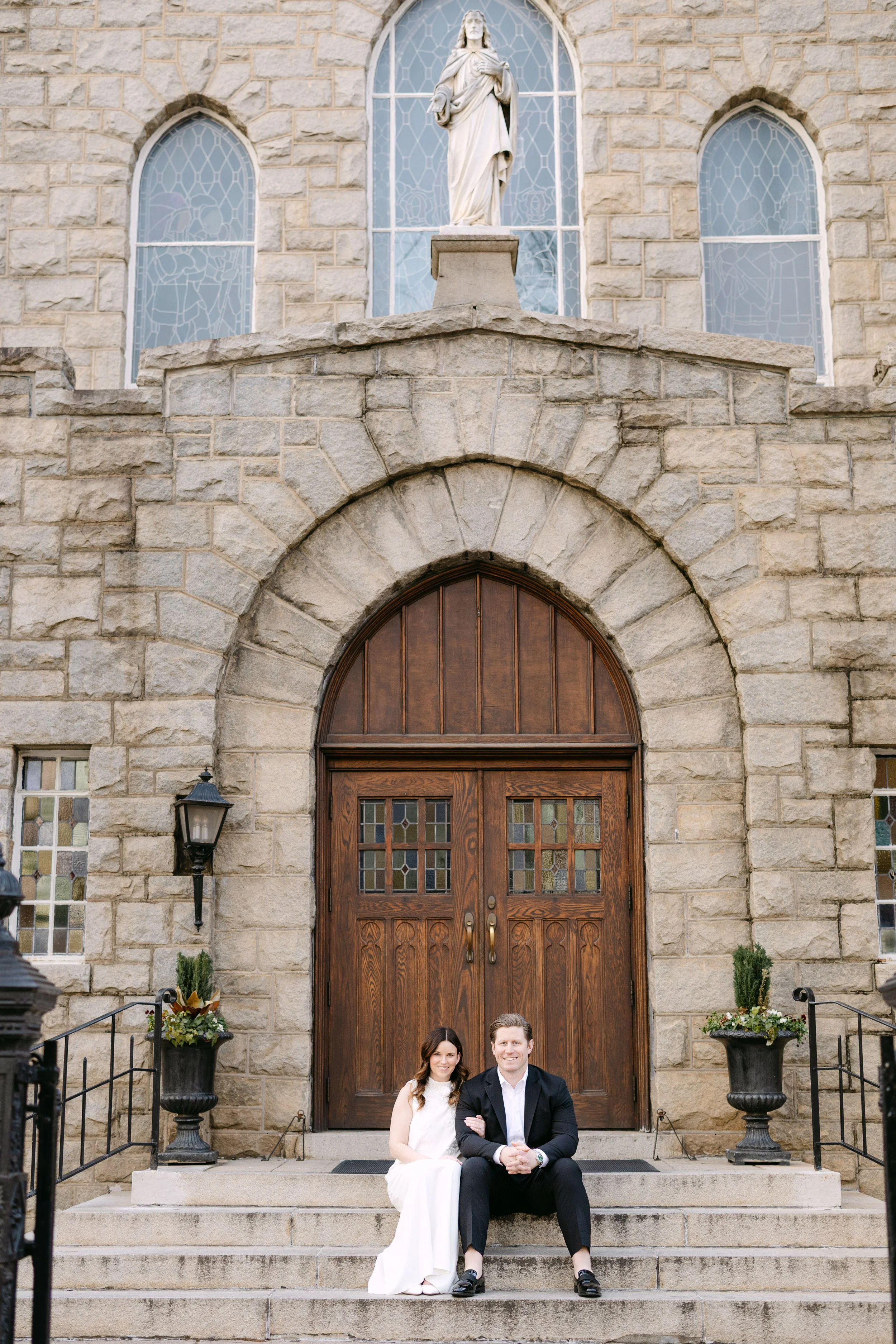 A couple dressed in formal attire sitting on stone steps in front of a large wooden church door, with stone walls, stained glass windows, and religious statues.
