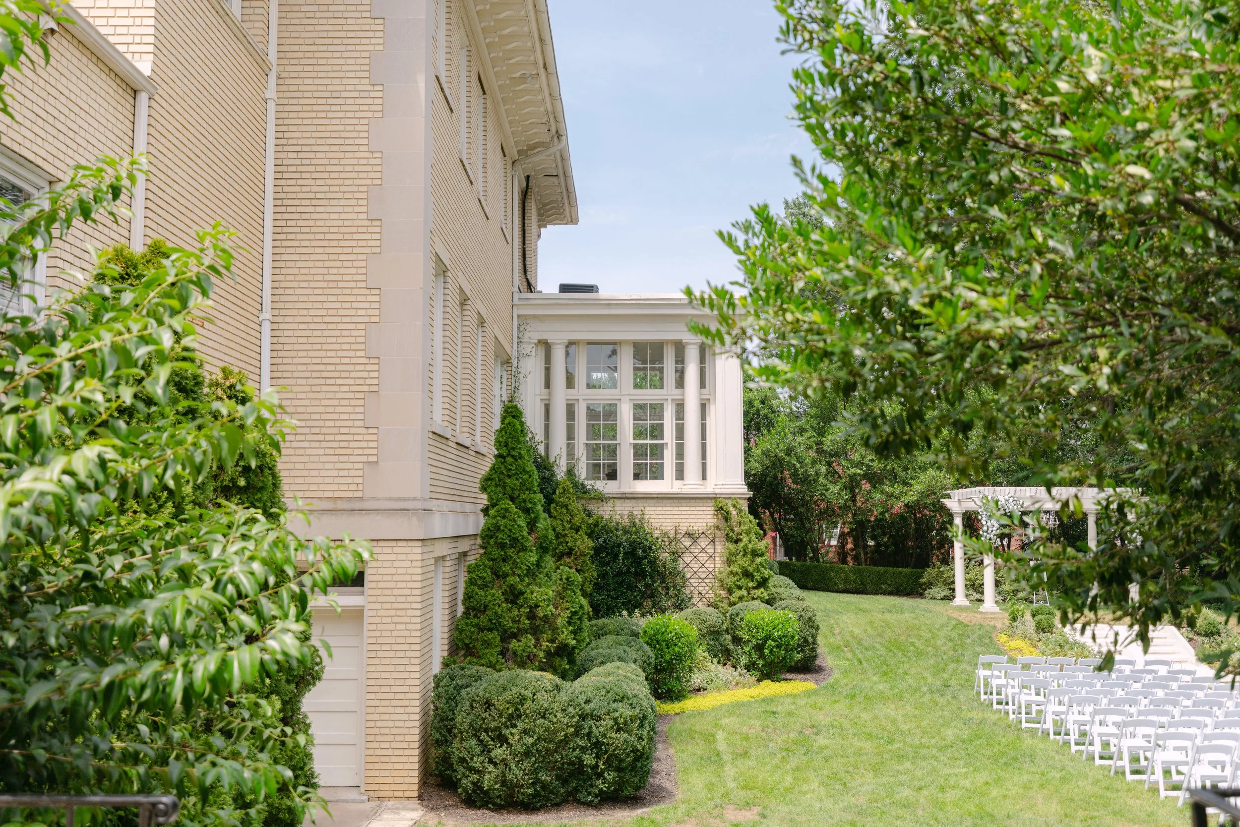 A well-maintained garden area with neatly trimmed bushes, green trees, and white chairs arranged outdoors, adjacent to a large beige brick house with white columns and windows, under a blue sky with some clouds.