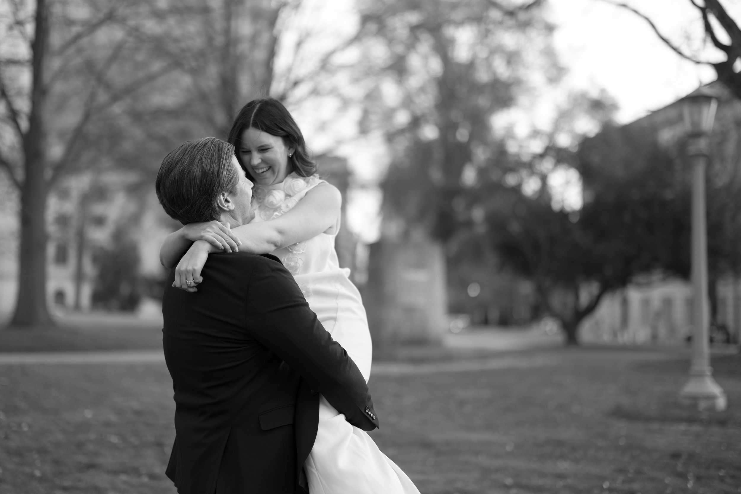 A man in a suit lifting a smiling woman in a white dress in a park, both looking happy.