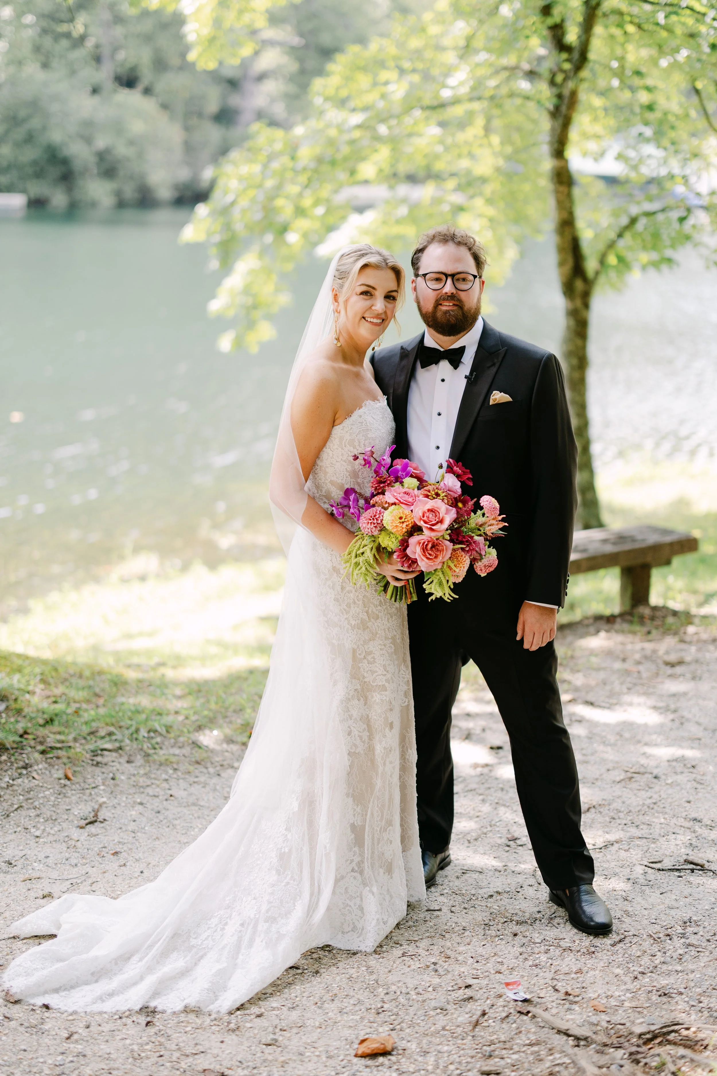 A bride and groom standing outdoors near a lake or river, with trees in the background. The bride is holding a colorful bouquet of pink, purple, and green flowers, and both are dressed in formal wedding attire.