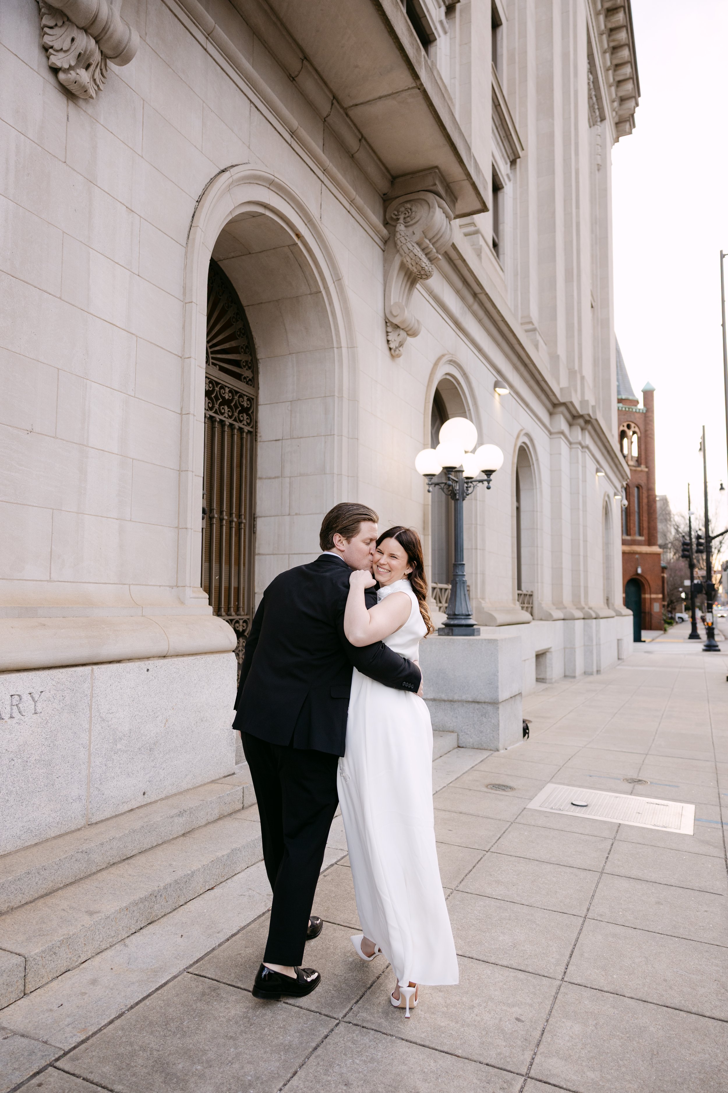 Couple dressed in wedding attire embracing and smiling outside a historic stone building with ornate architectural details and street lamps.