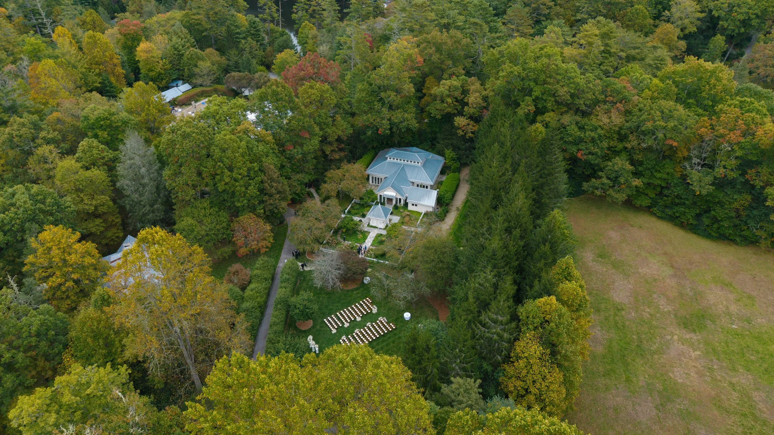 Aerial view of a gray-roofed building surrounded by colorful autumn forest, gardens, pathways, and rows of outdoor seating.