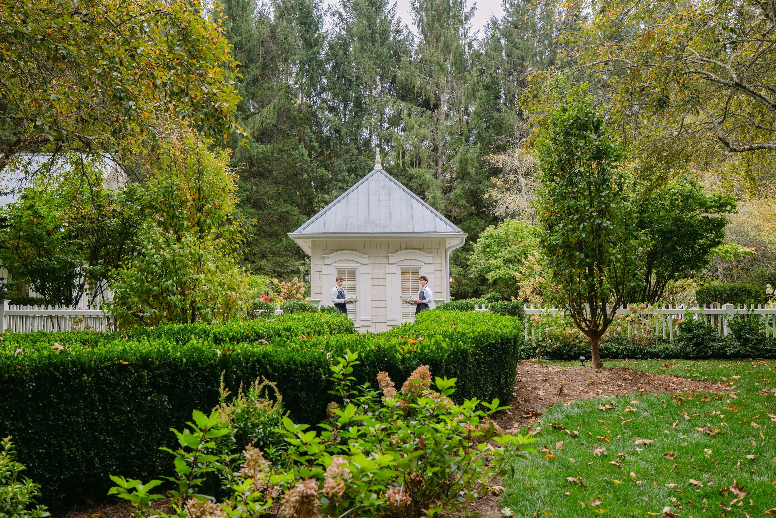 Two servers stand outside a small white garden pavilion surrounded by manicured hedges and autumn foliage.
