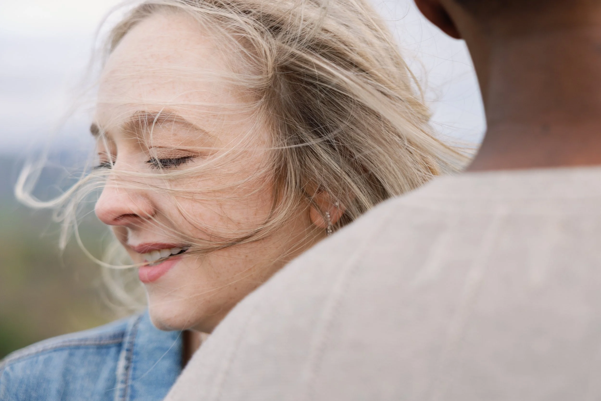 A young woman with blonde hair smiling, with wind blowing through her hair, wearing a denim jacket.