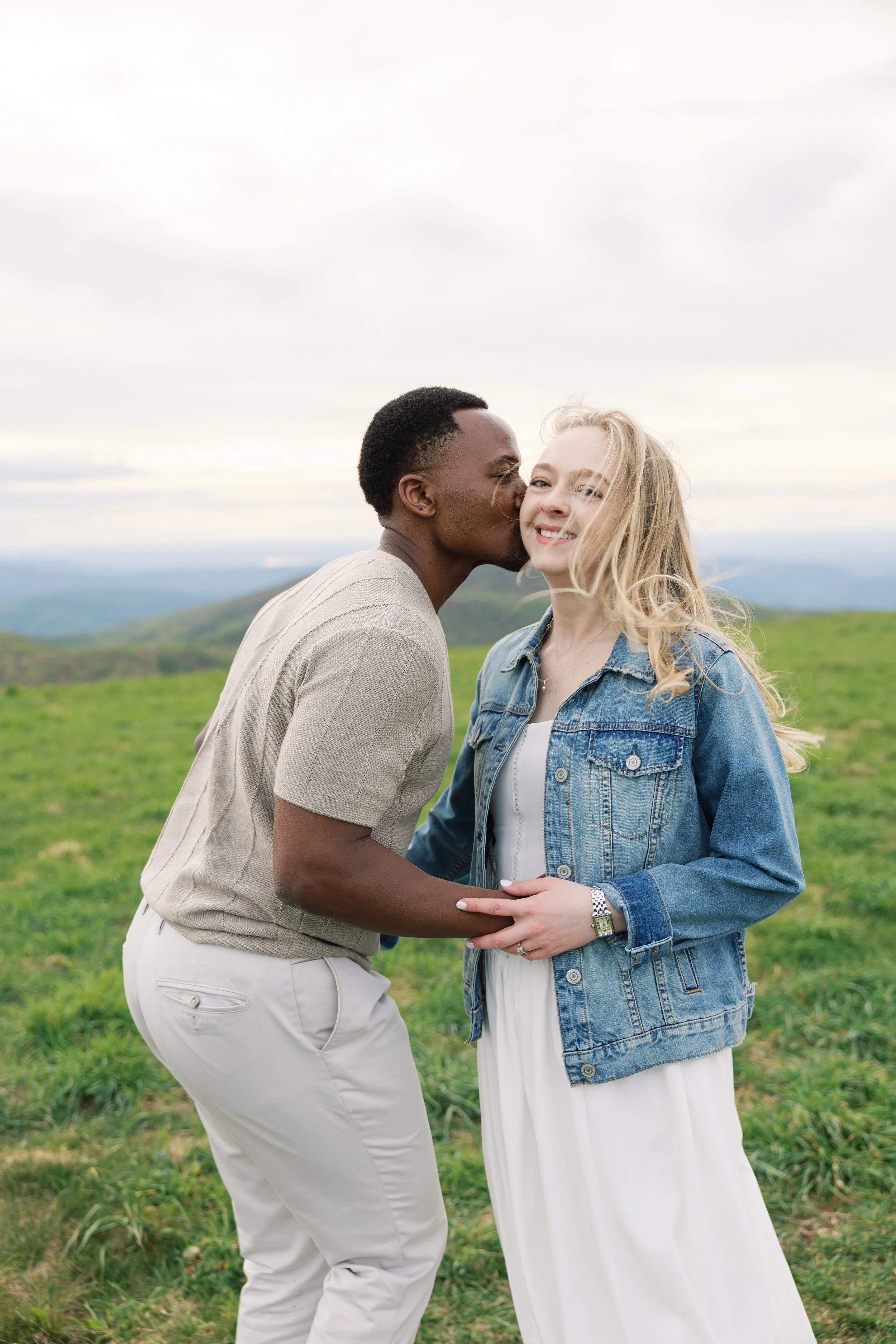 A couple sharing an outdoor moment, with the man kissing the woman on the cheek during daytime in a field, wearing casual clothing, with a landscape of green hills and cloudy sky in the background.
