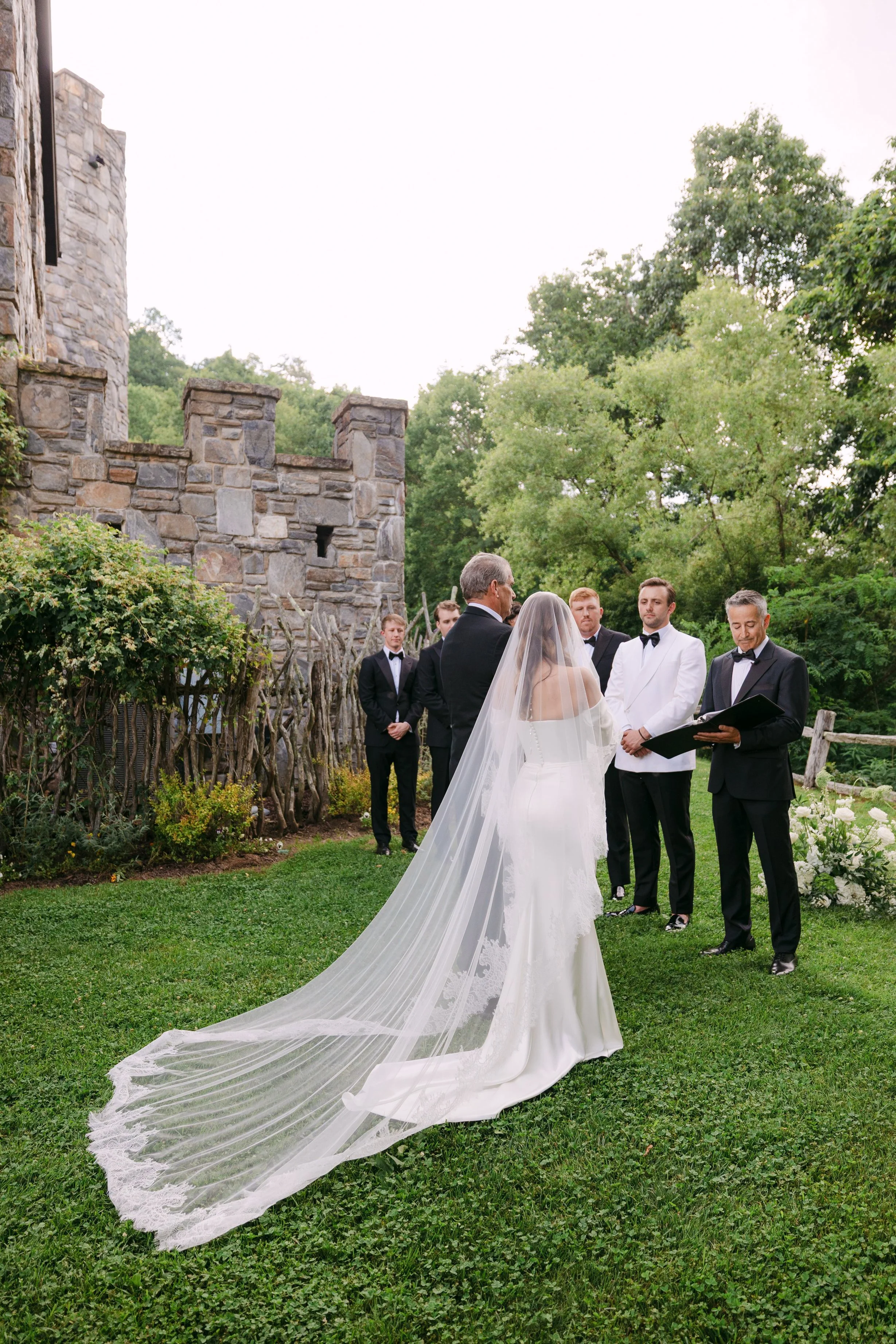 Bride in a white wedding dress with a long veil facing an officiant and five groomsmen in formal black and white tuxedos, outside near a stone building and greenery, during a wedding ceremony.