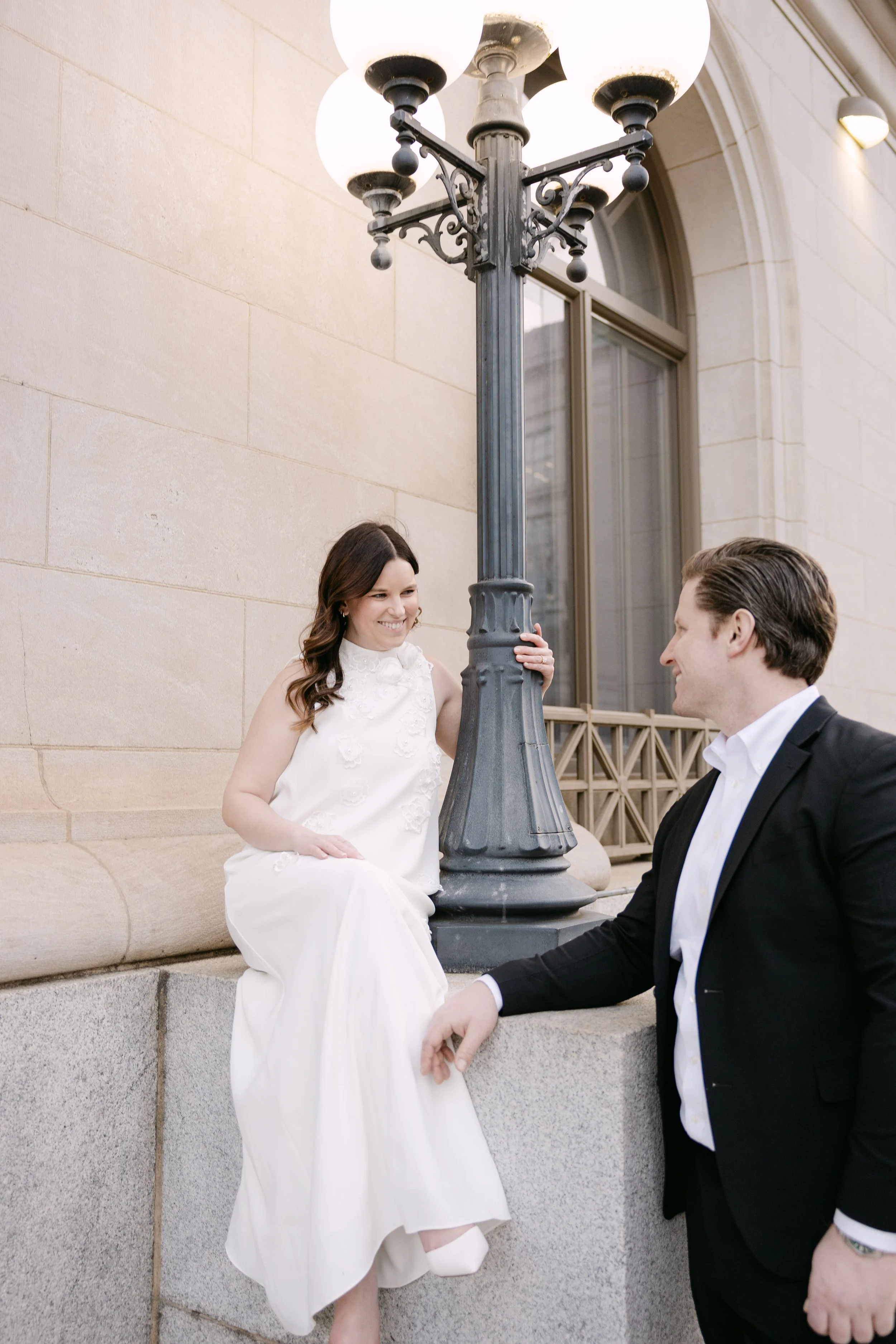 A woman in a white dress and a man in a black suit share a moment near a vintage street lamp outside a building with beige stone walls.