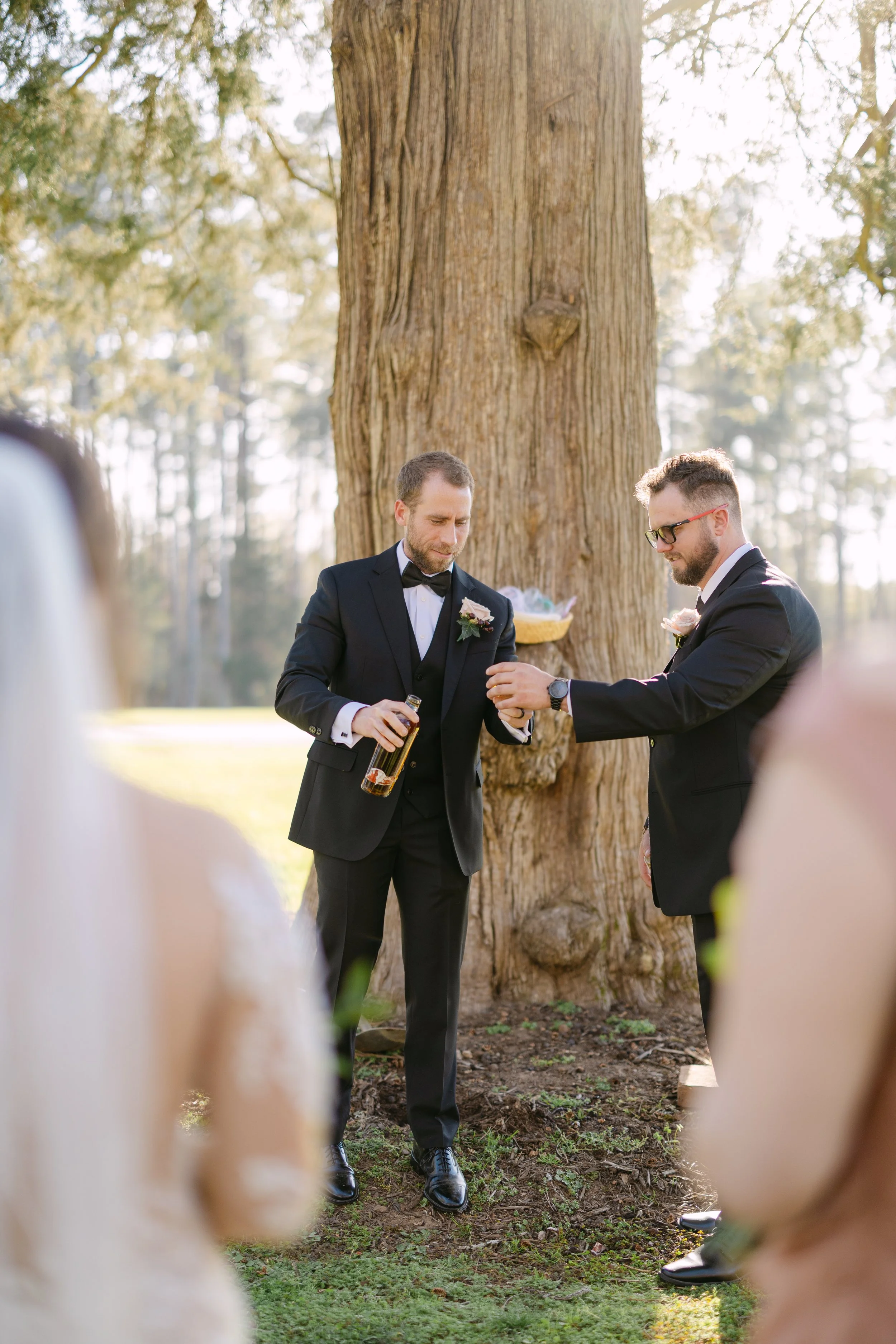 Two men in tuxedos, one holding a bottle, stand outdoors near a large tree, engaging in a wedding ceremony with blurred guests in the foreground.
