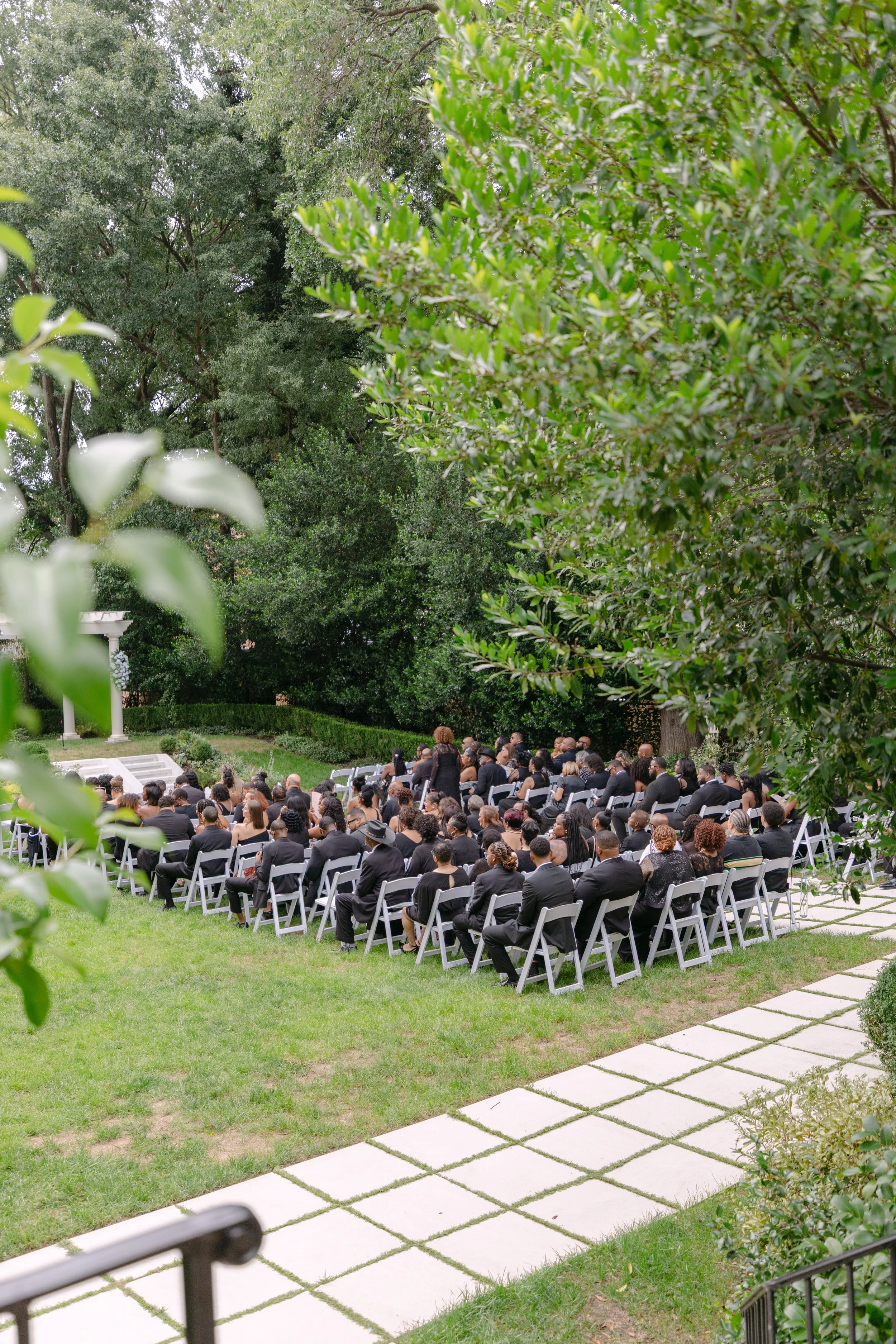 Outdoor event with seated attendees dressed in black, facing a speaker at a podium in a garden surrounded by lush green trees.