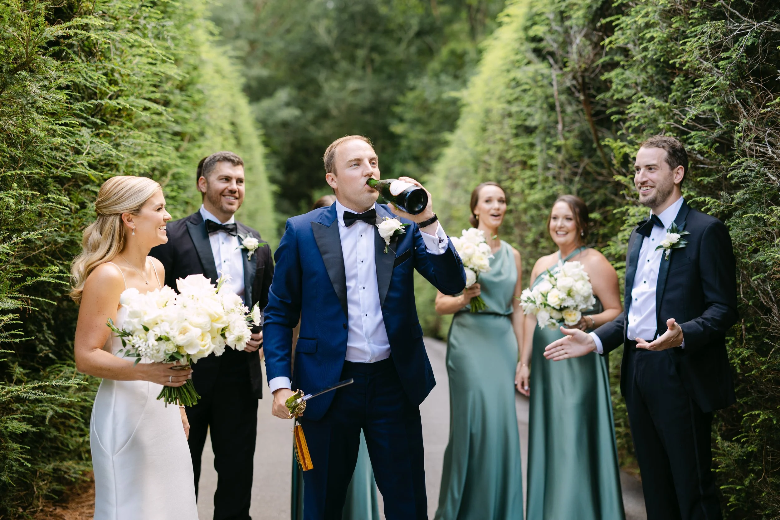 Group of wedding guests dressed in formal attire, standing outdoors on a lush green path, with one man drinking from a bottle while others smile and chat.