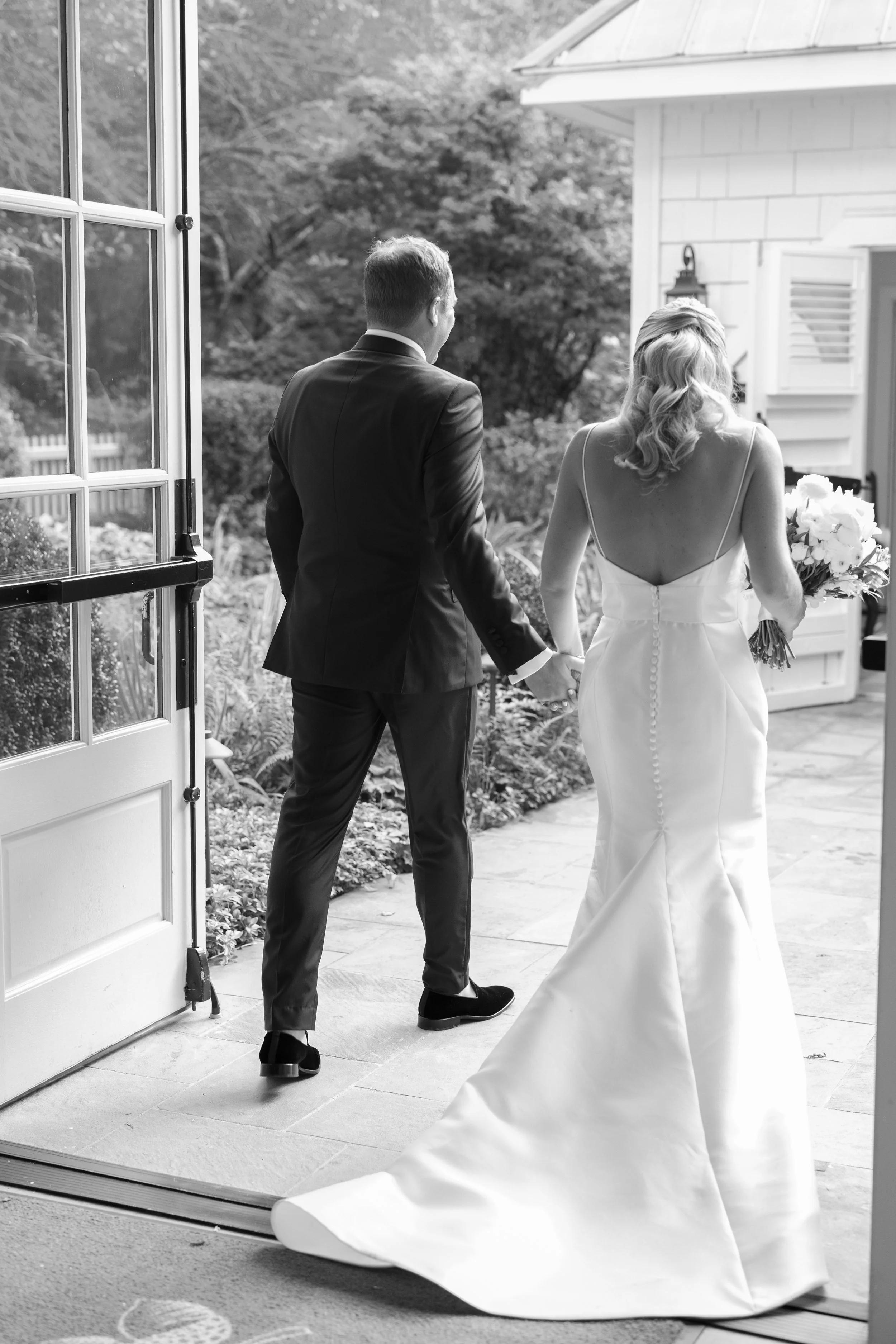 A bride in a wedding dress holding a bouquet and a groom in a suit walking hand-in-hand out of a house.