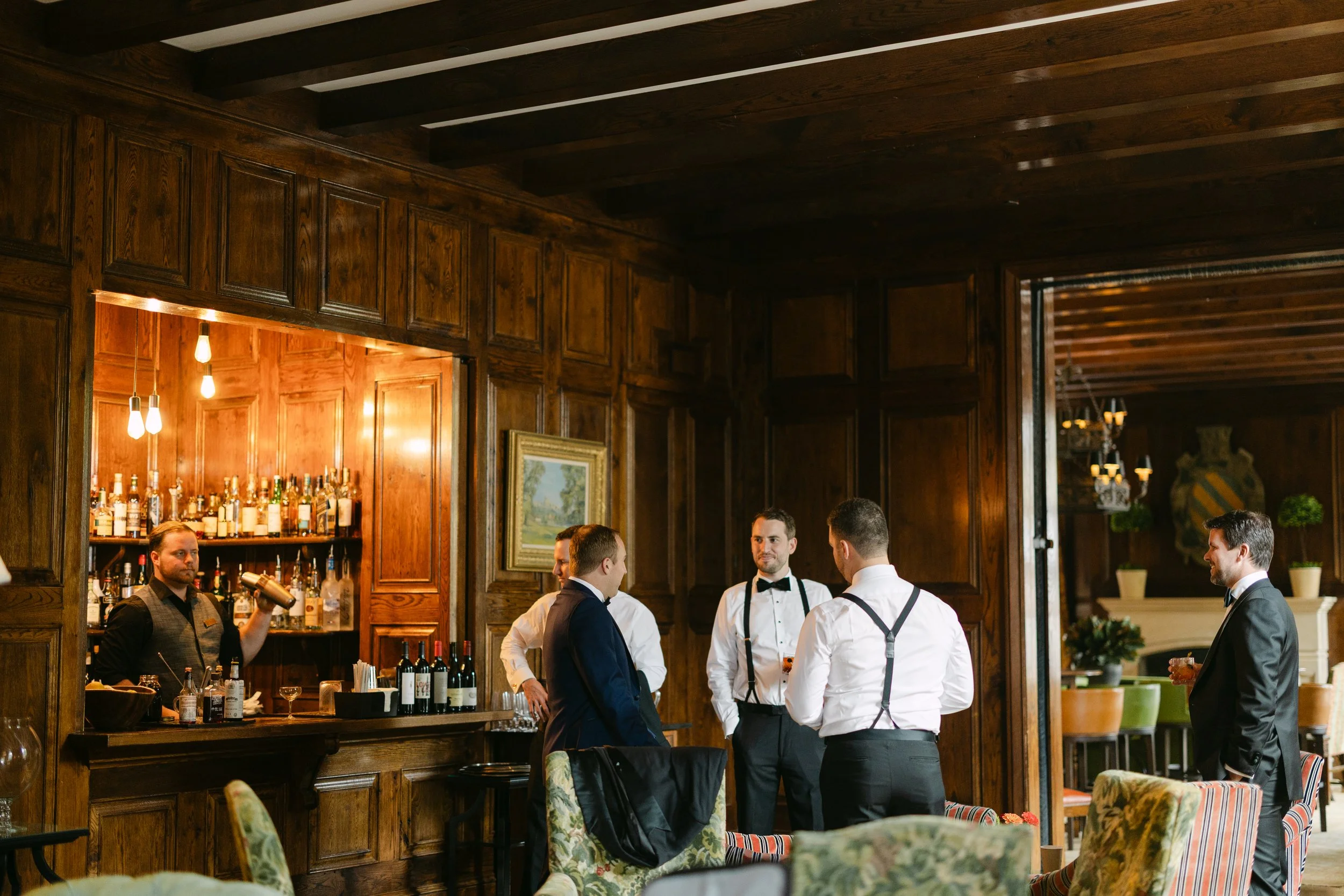 Group of five men in suits and suspenders talking in a formal lounge. The bar area with bottles and bartender in the background, wood-paneled walls, and elegant decor.