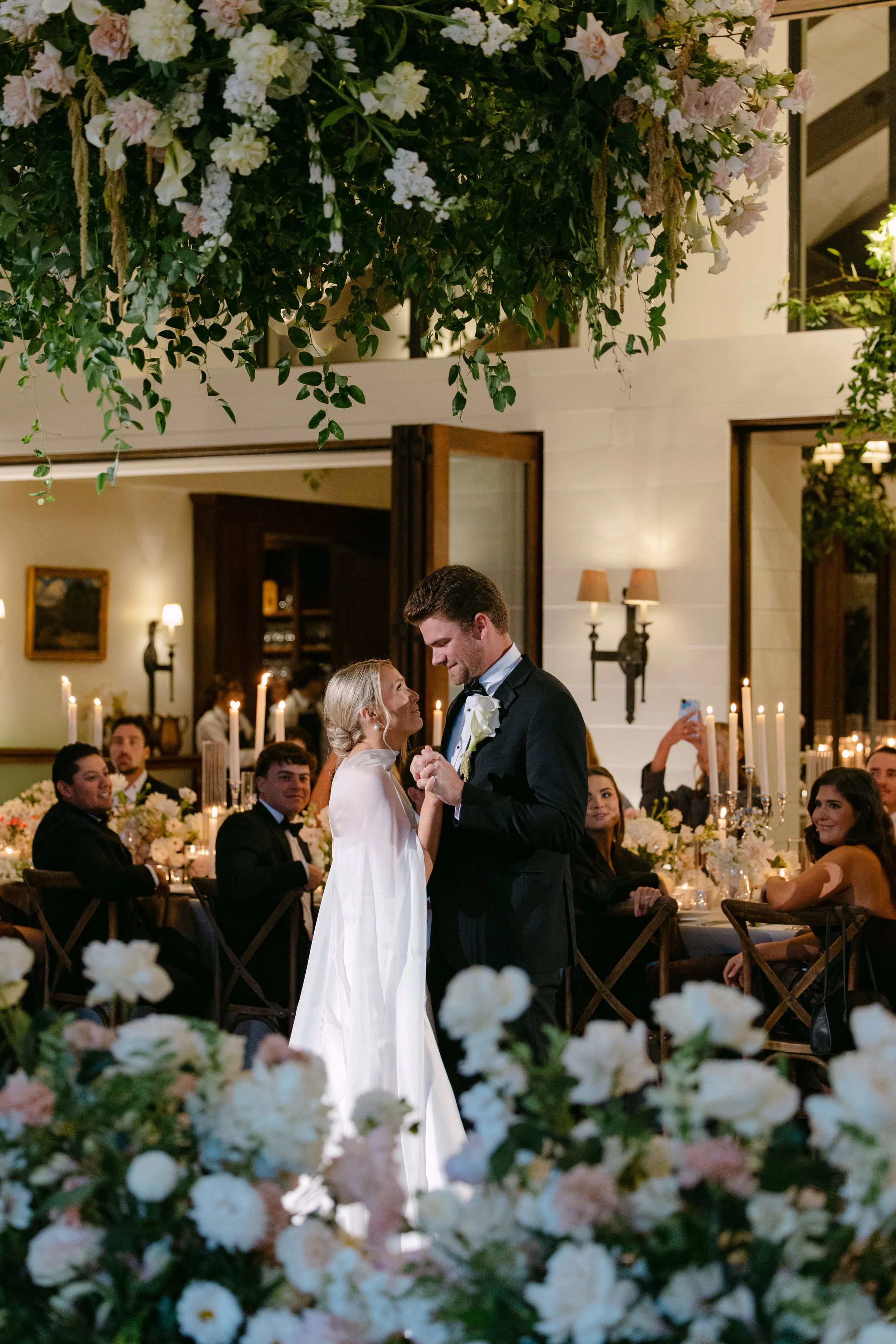 Bride and groom share a first dance at a candlelit wedding reception surrounded by lush white and blush floral arrangements.