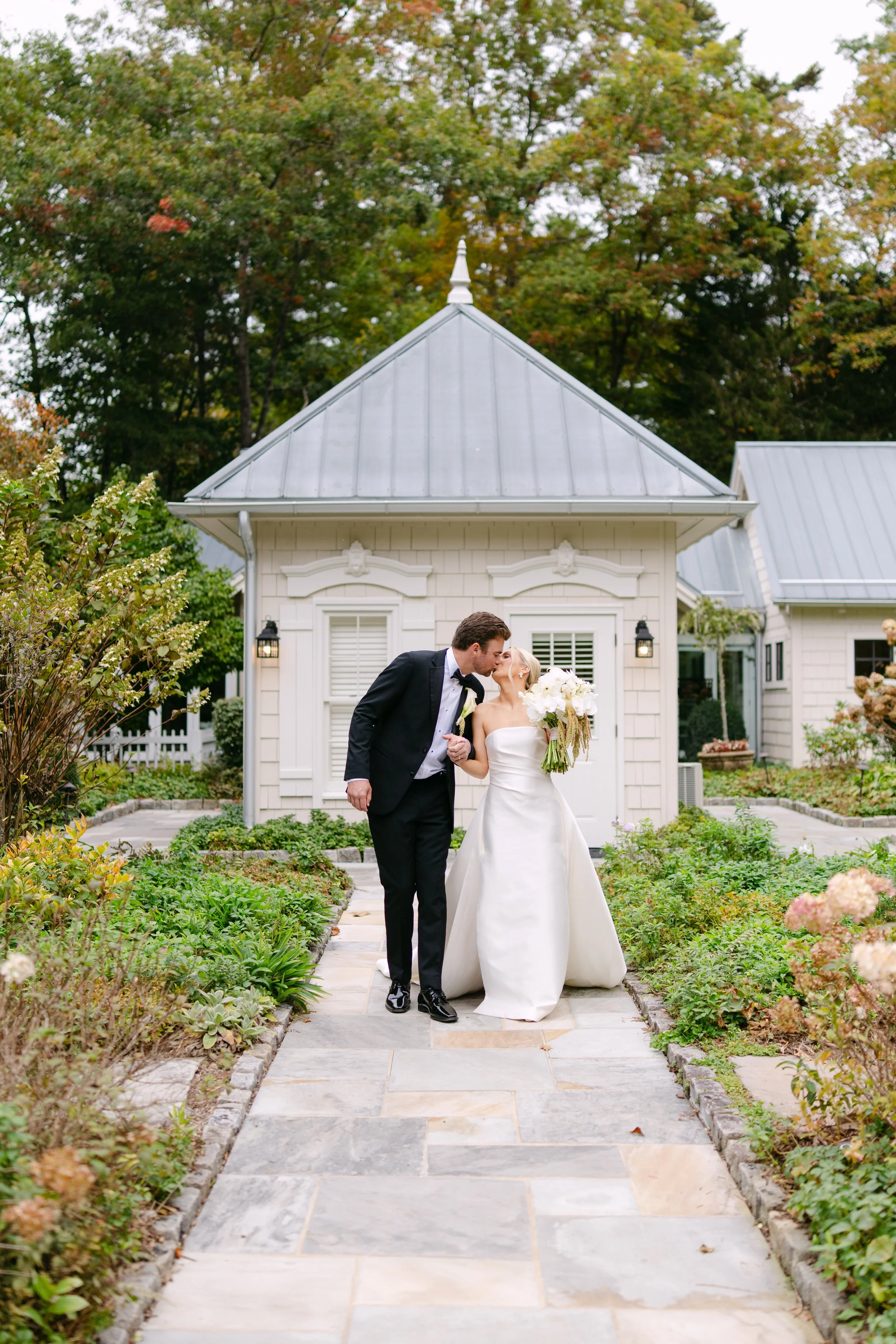 Newlywed couple kissing outside a white wedding venue, with the bride holding a bouquet of white flowers.