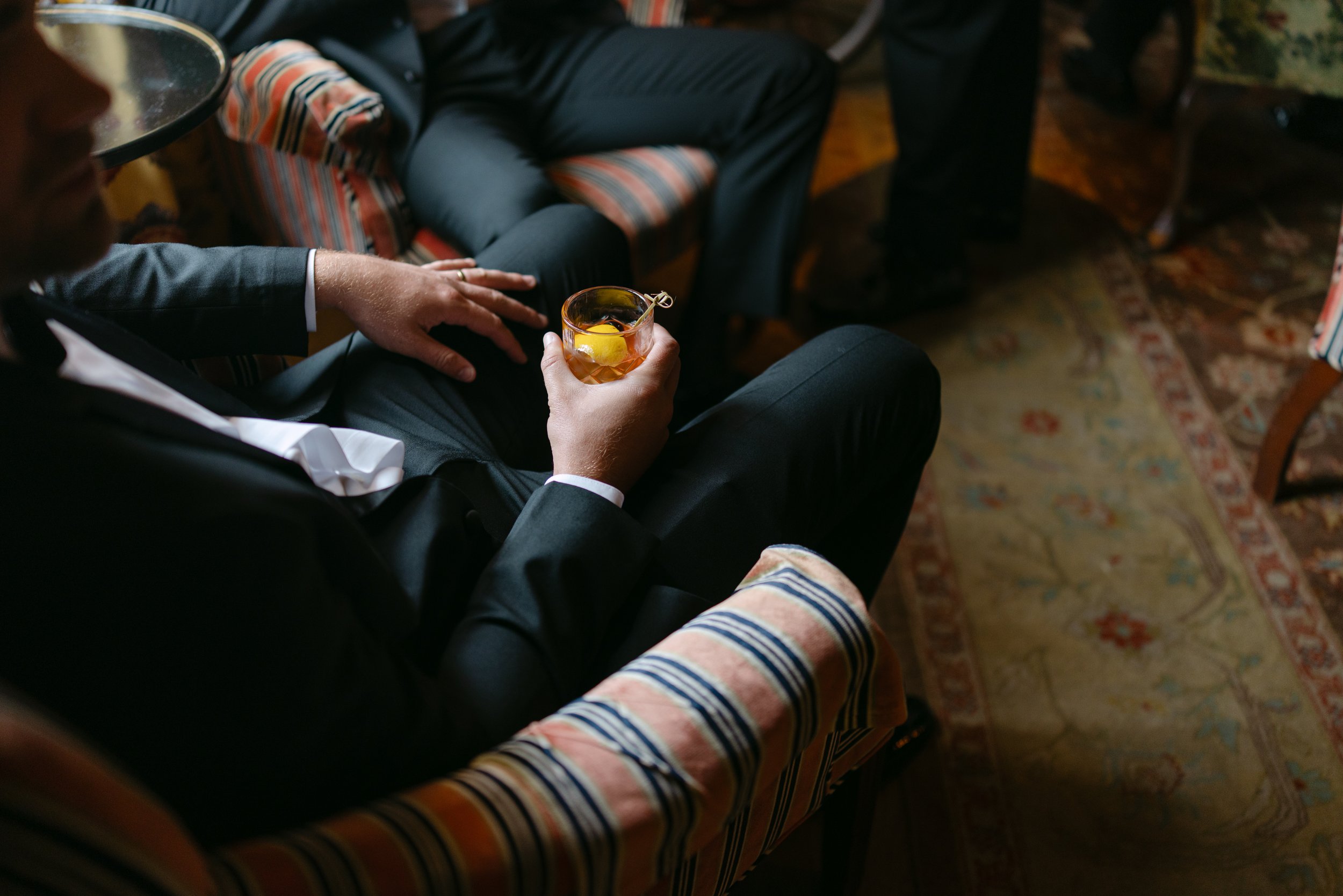 Man in a dark suit holding a whiskey cocktail with a lemon peel while seated in a striped armchair