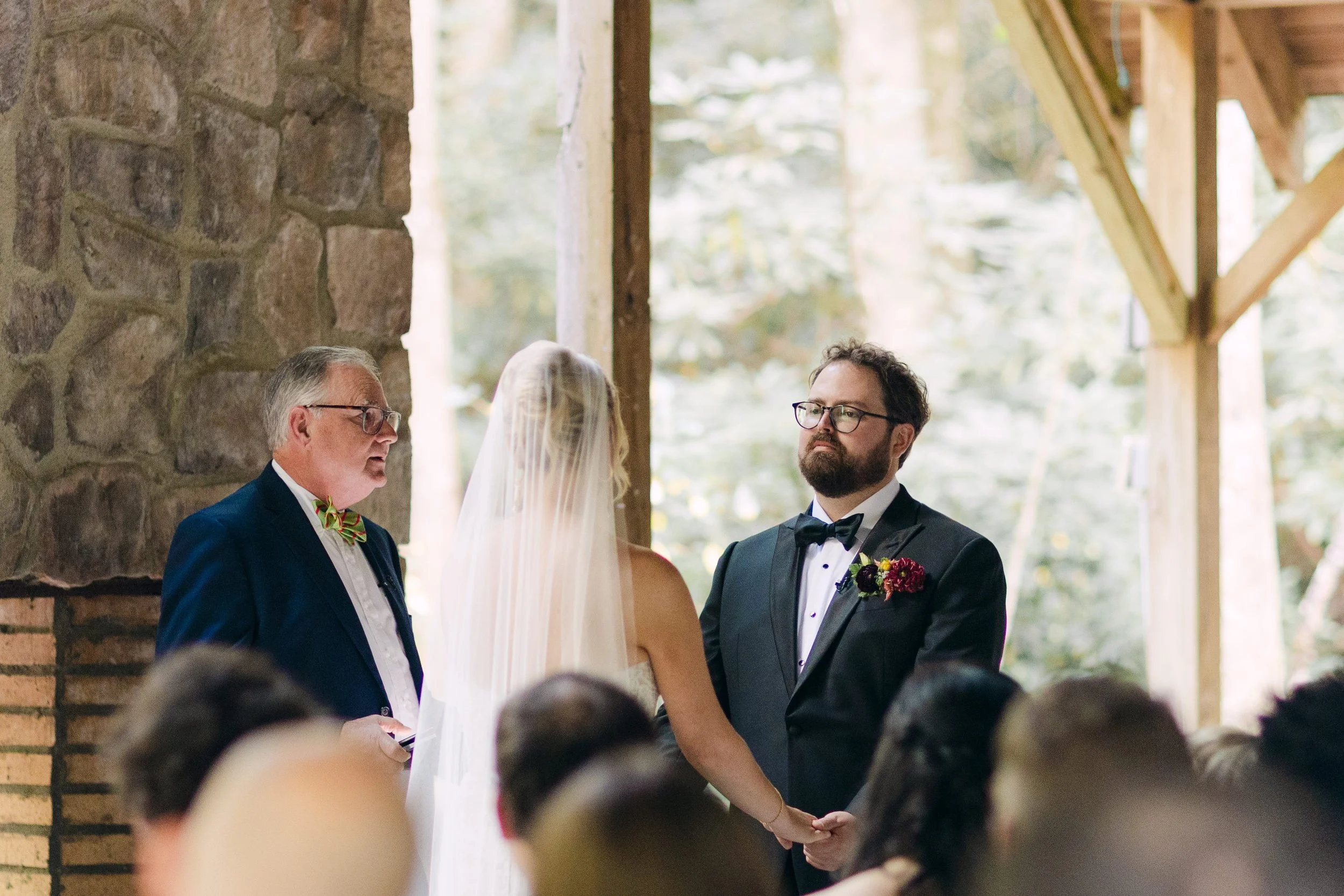 A wedding ceremony with a bride and groom holding hands, facing a officiant inside a rustic venue with stone walls and wooden beams.