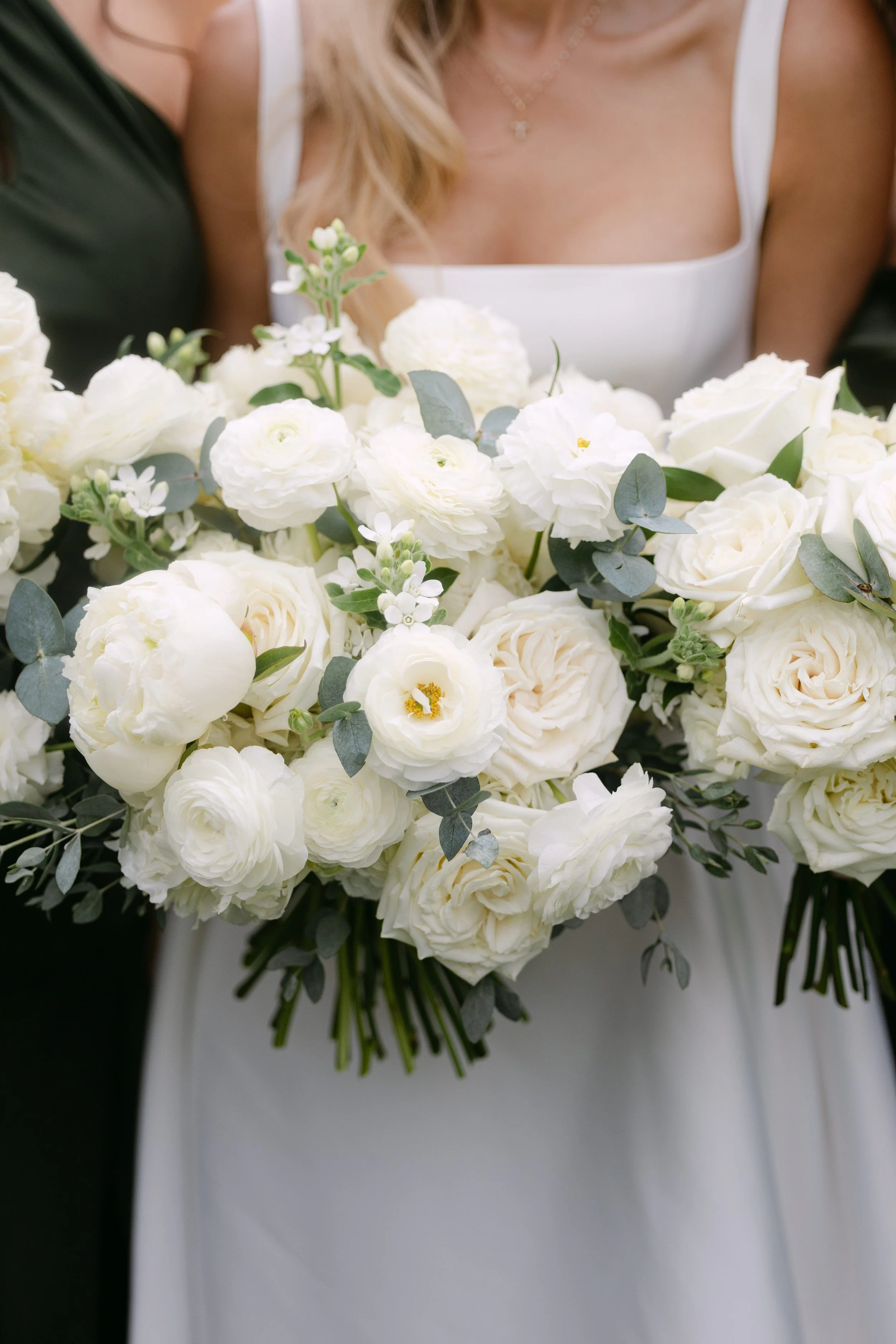 Bride holding a large bouquet of white flowers, including roses and peonies, with greenery, at a wedding.