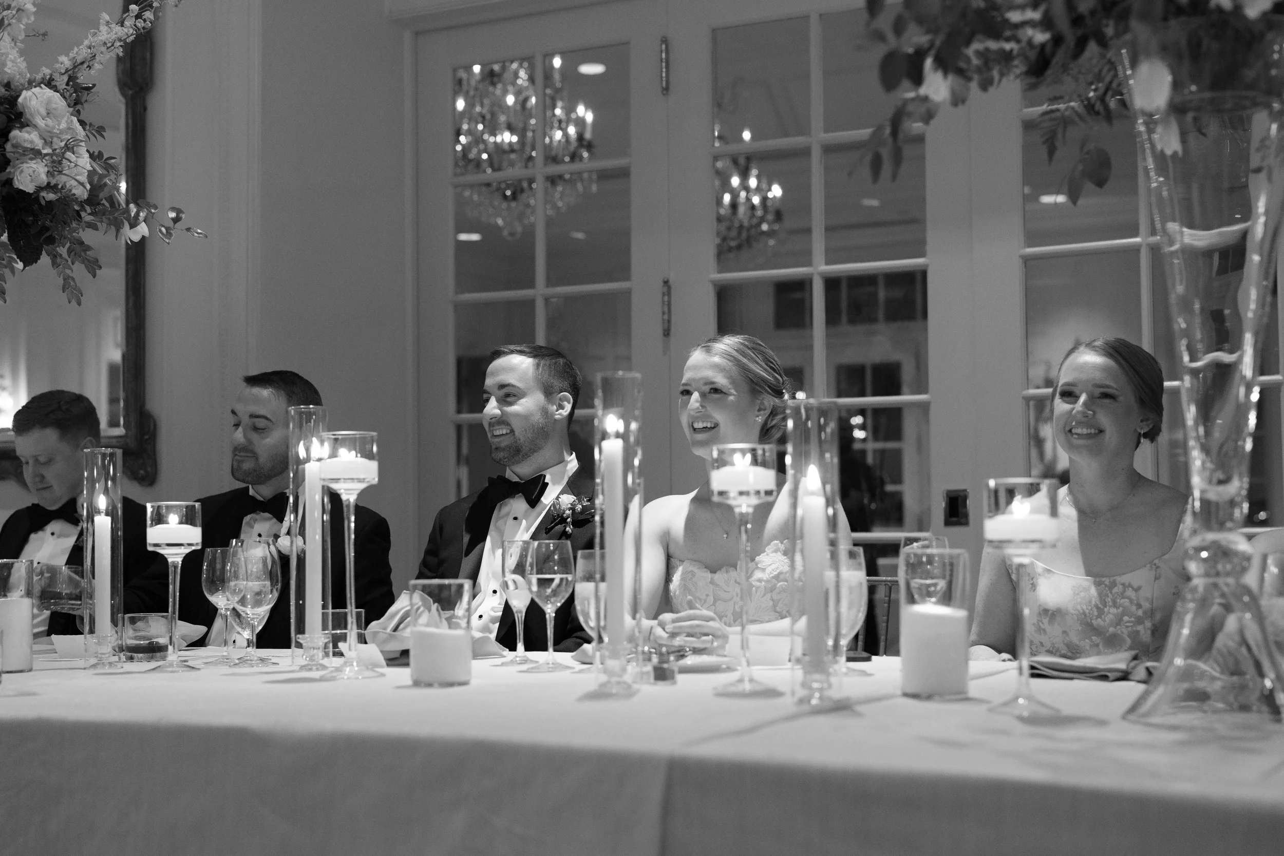A black and white photograph of a wedding reception with five people seated at a decorated head table, including the bride and groom, sitting between two bridesmaids and two groomsmen, surrounded by candles and floral arrangements.