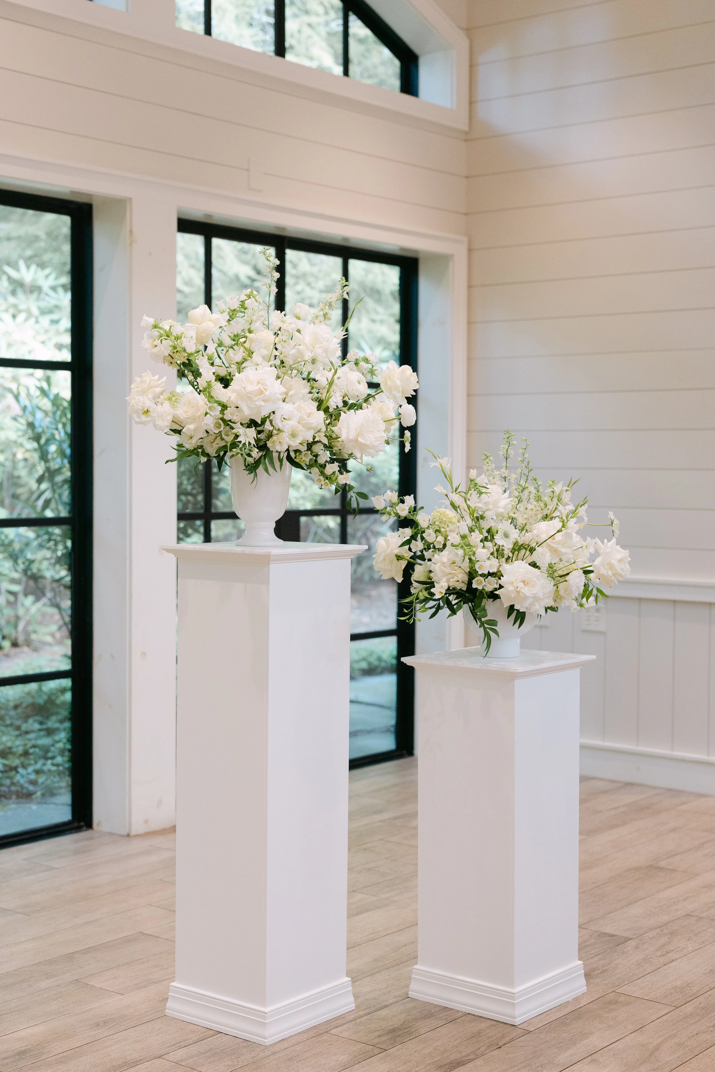 Two white floral arrangements in vases on white pedestals inside a room with large windows and wooden flooring.