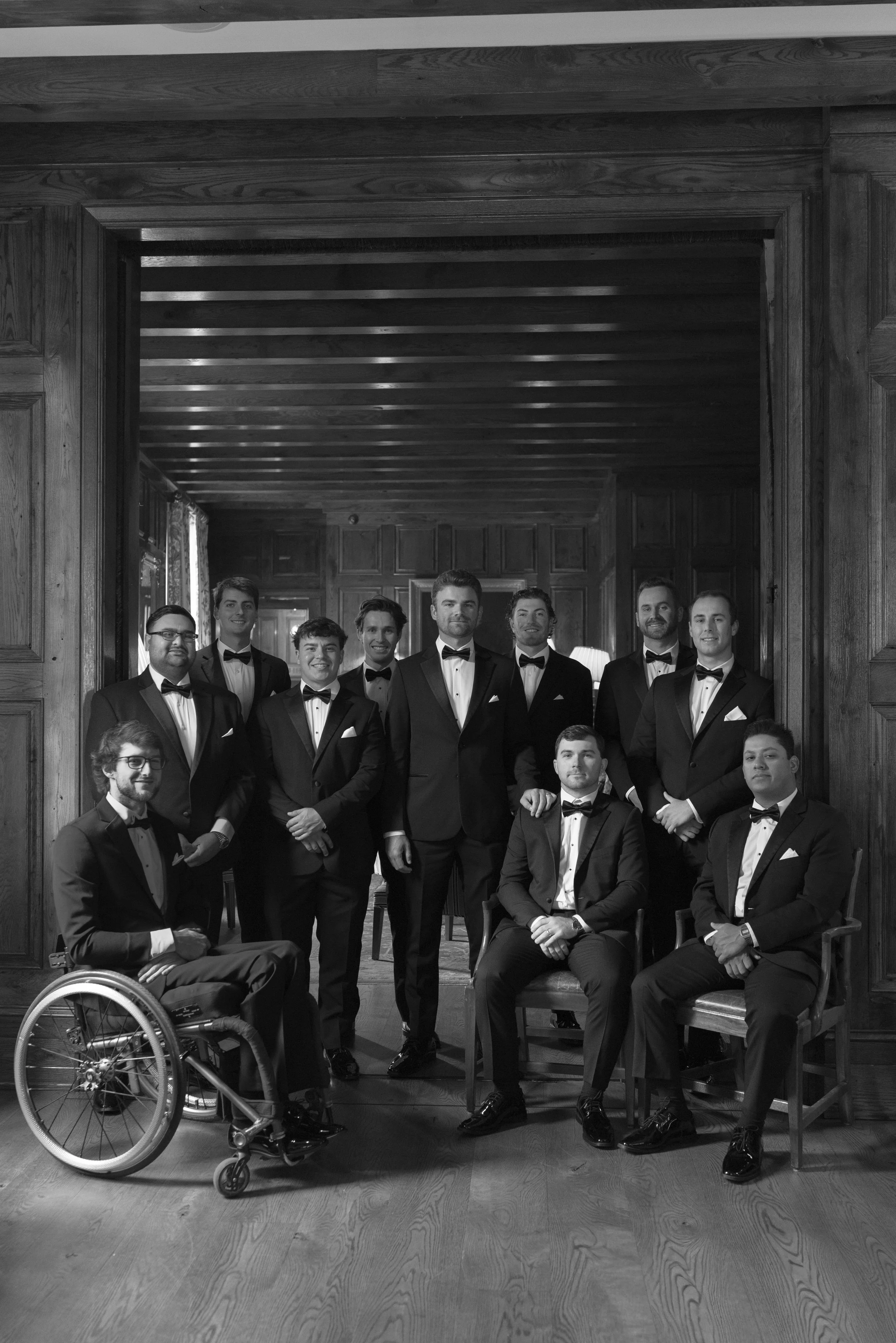 Black-and-white group portrait of men in tuxedos, including one man in a wheelchair, posing inside a wood-paneled room.