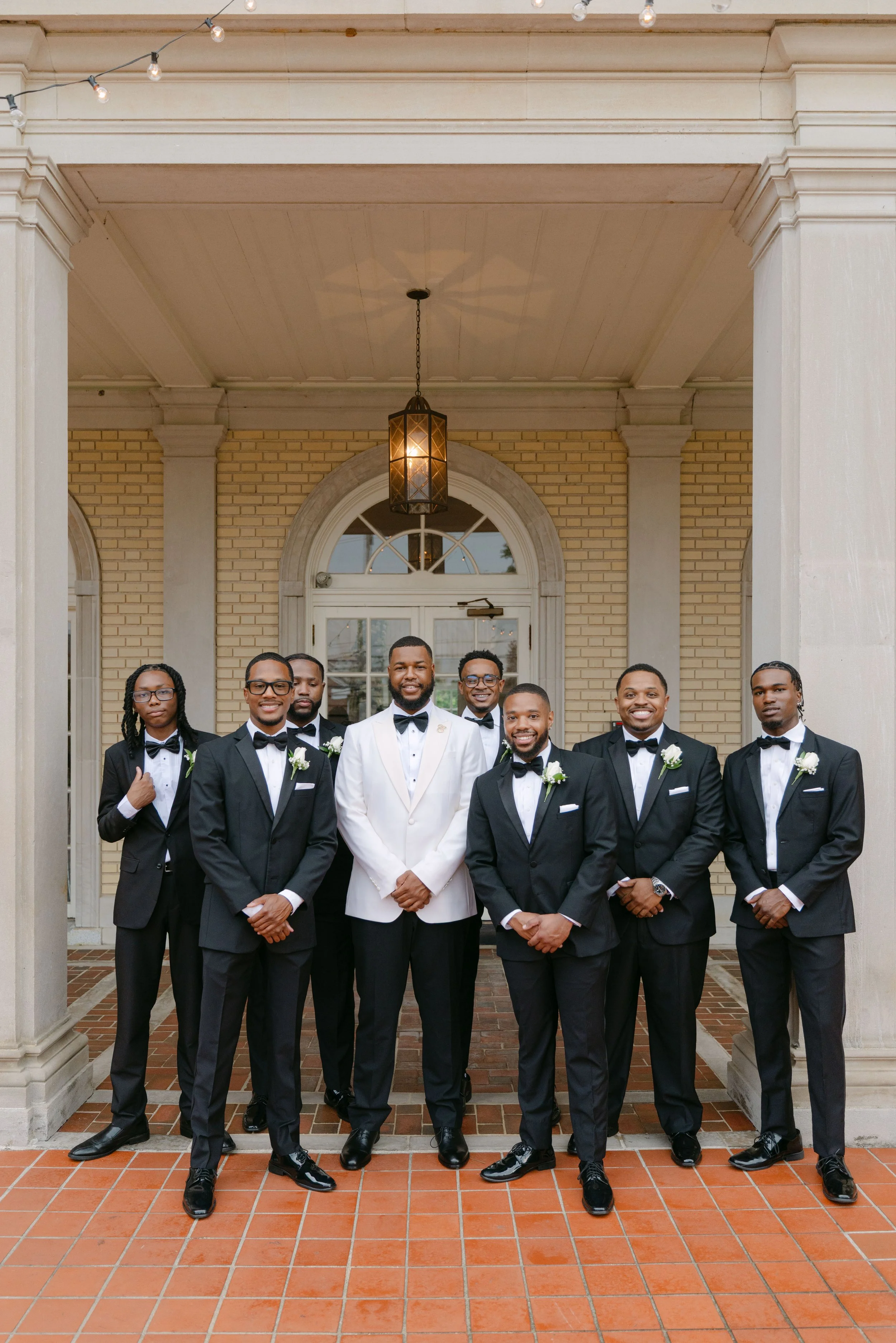 Group of men dressed in black tuxedos with white shirts and black bow ties standing outside in front of a brick building with a lantern hanging from the ceiling. The group is smiling and looking at the camera.