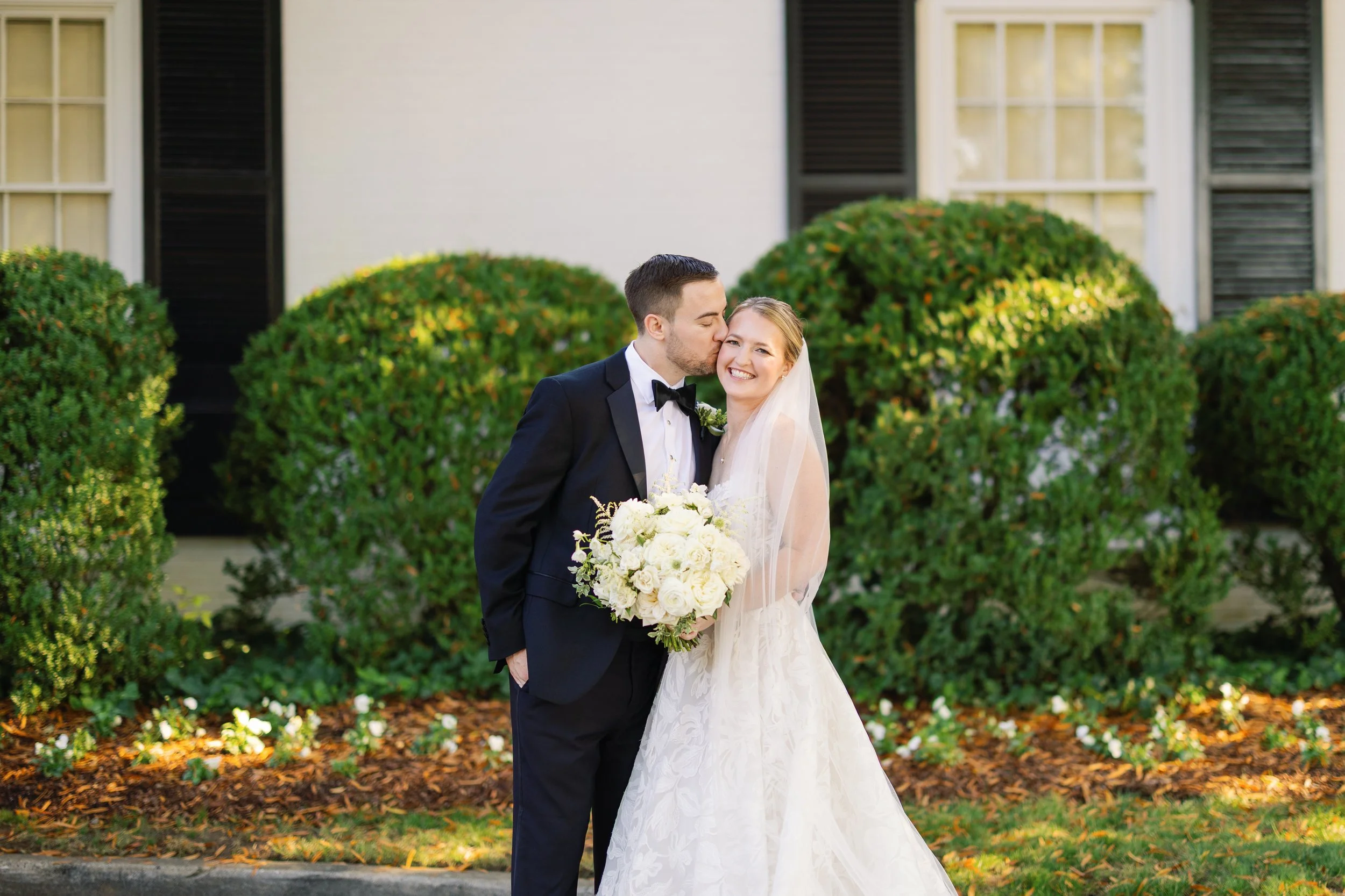 A bride and groom standing outdoors, with the groom kissing the bride's temple while she smiles at the camera. The bride is holding a large bouquet of white roses, and the groom is dressed in a black tuxedo with a bow tie. Behind them are green bushe