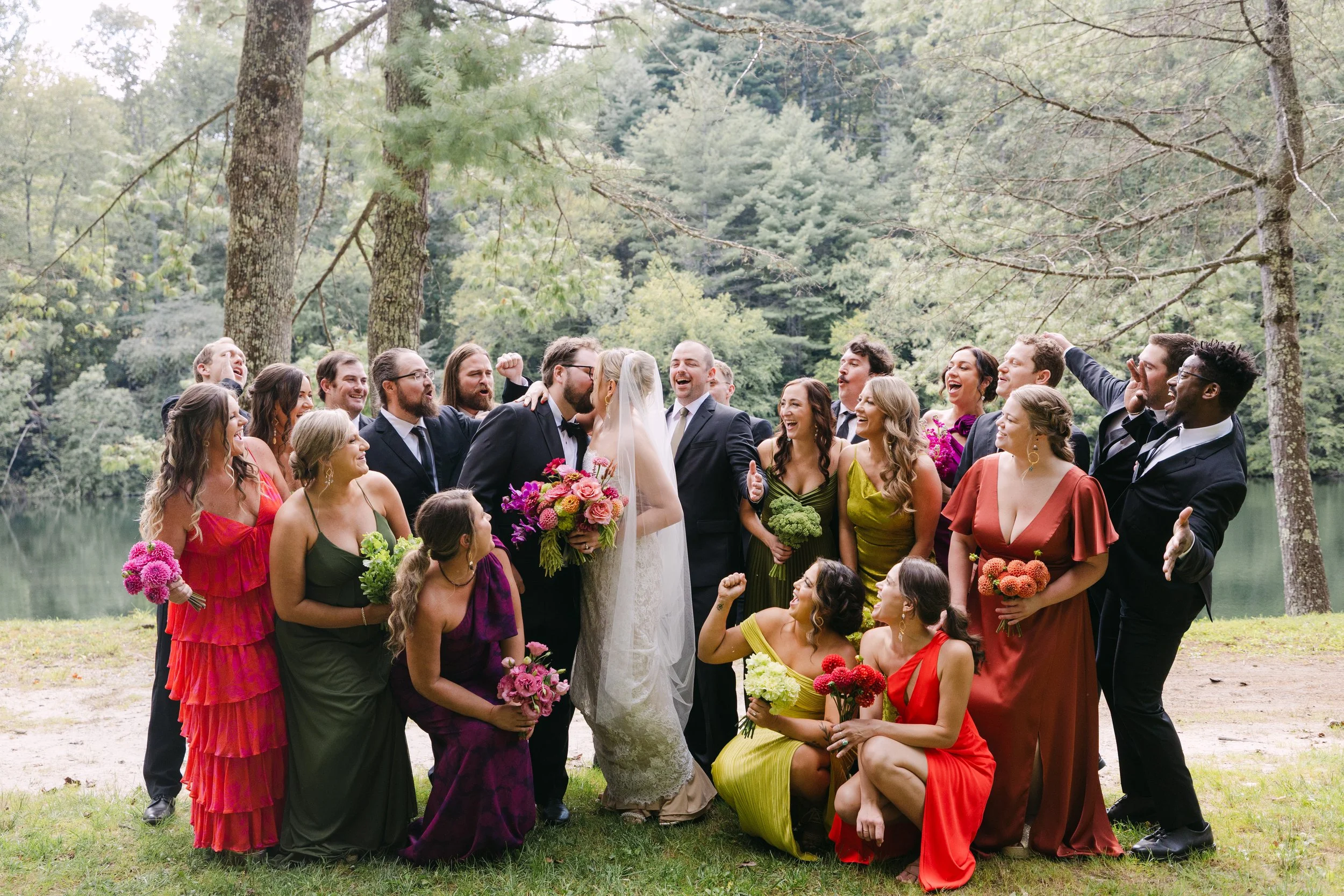 Group of wedding guests with a bride and groom outdoors near a lake, surrounded by trees, celebrating and smiling.