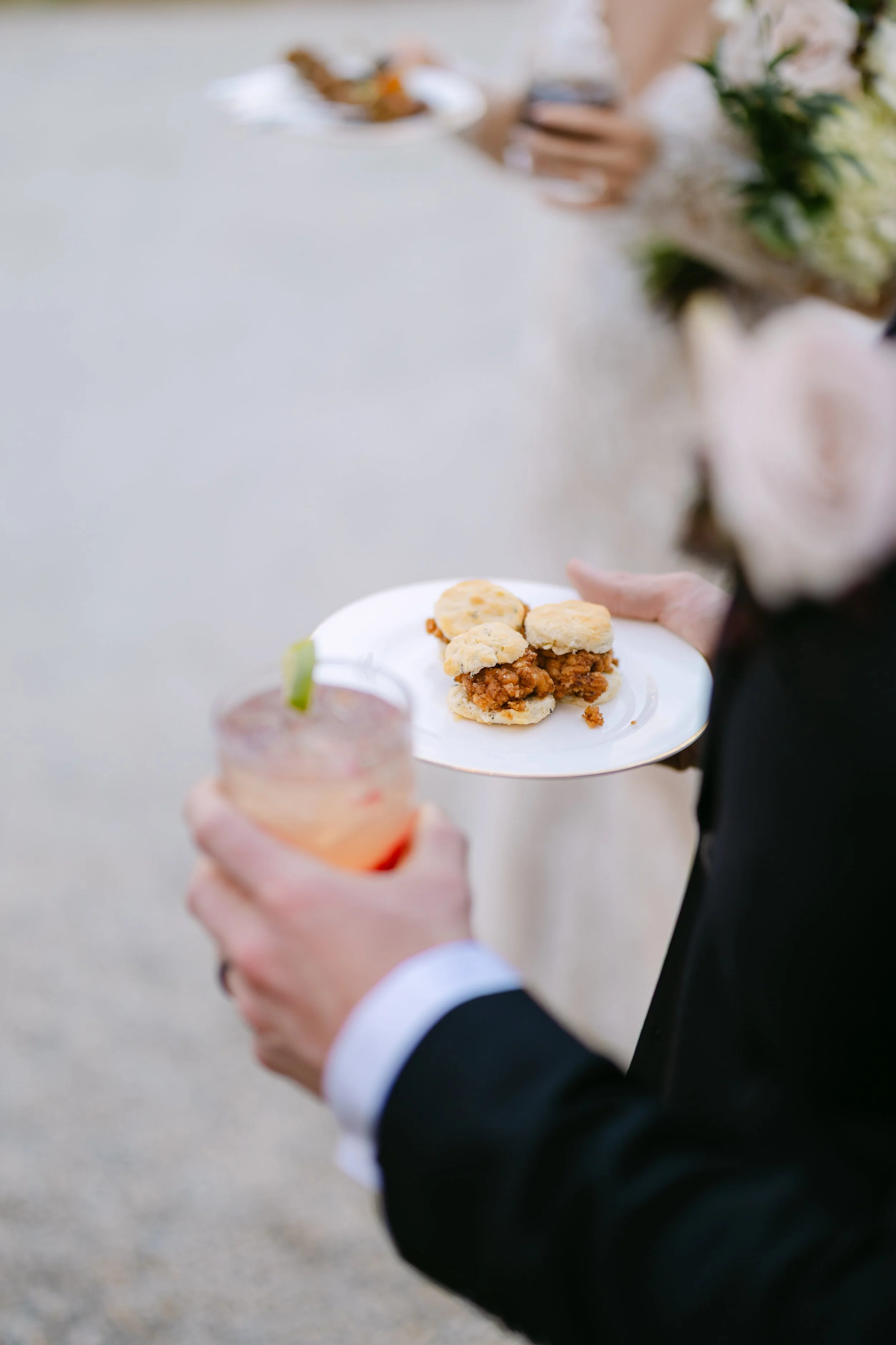 Person holding a plate with mini sandwiches and a drink with lime wedge at a social event.
