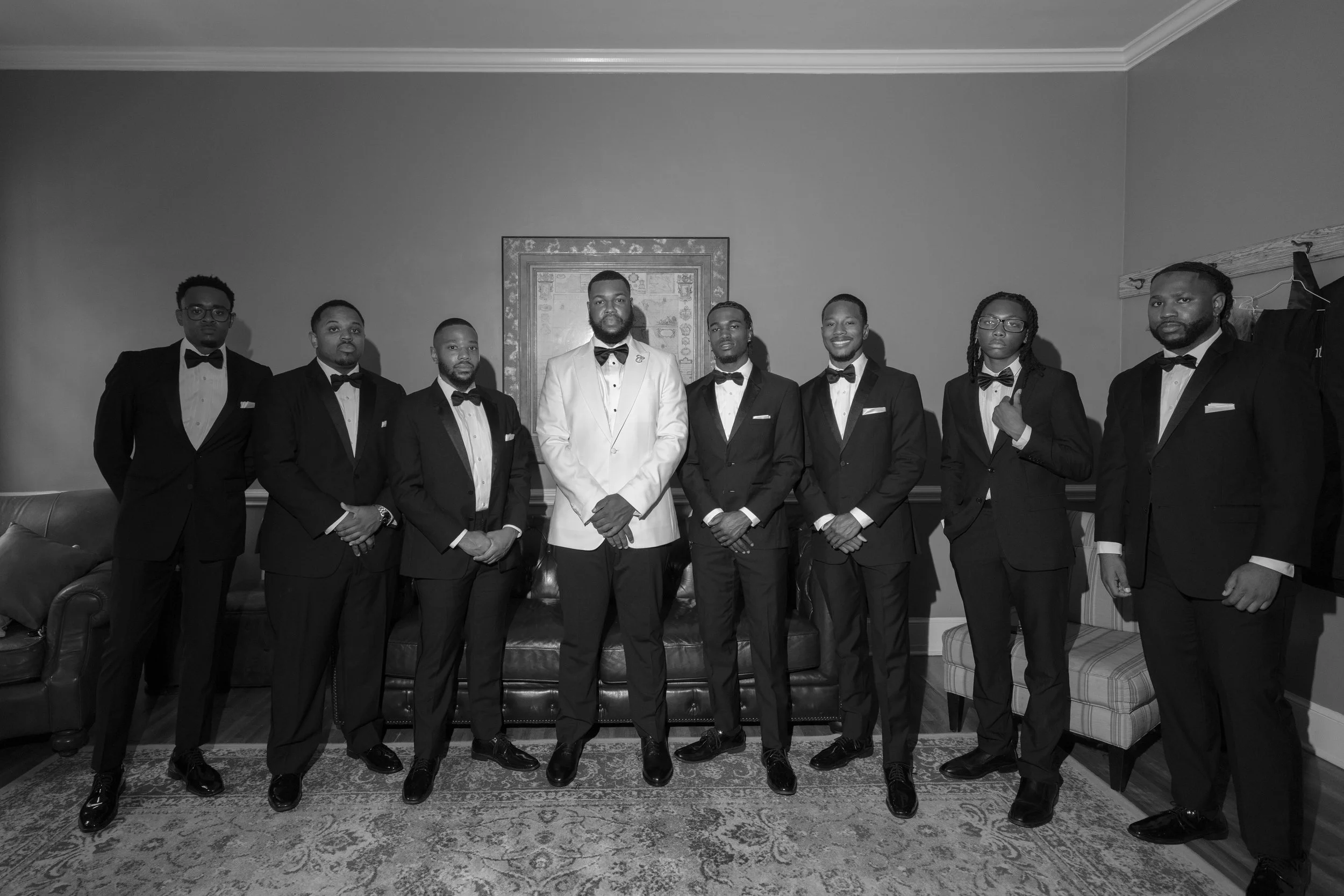 A group of nine men dressed in tuxedos, standing indoors against a wall with a framed picture. The central man is wearing a white tuxedo, while the others are in dark tuxedos, all with bow ties.